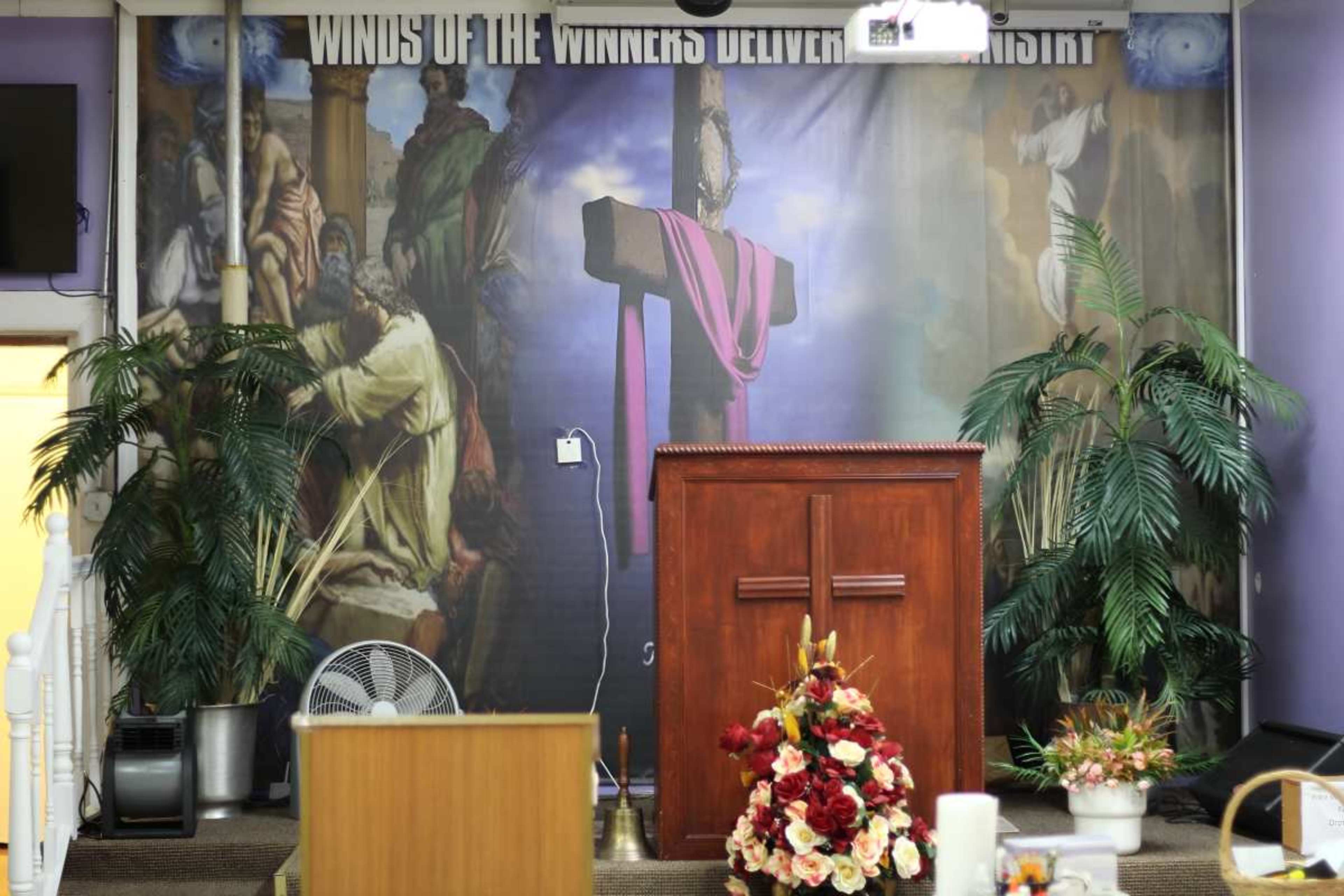 The image shows a church interior with a wooden podium in front of a large mural depicting biblical scenes and a cross, framed by potted plants and a fan.