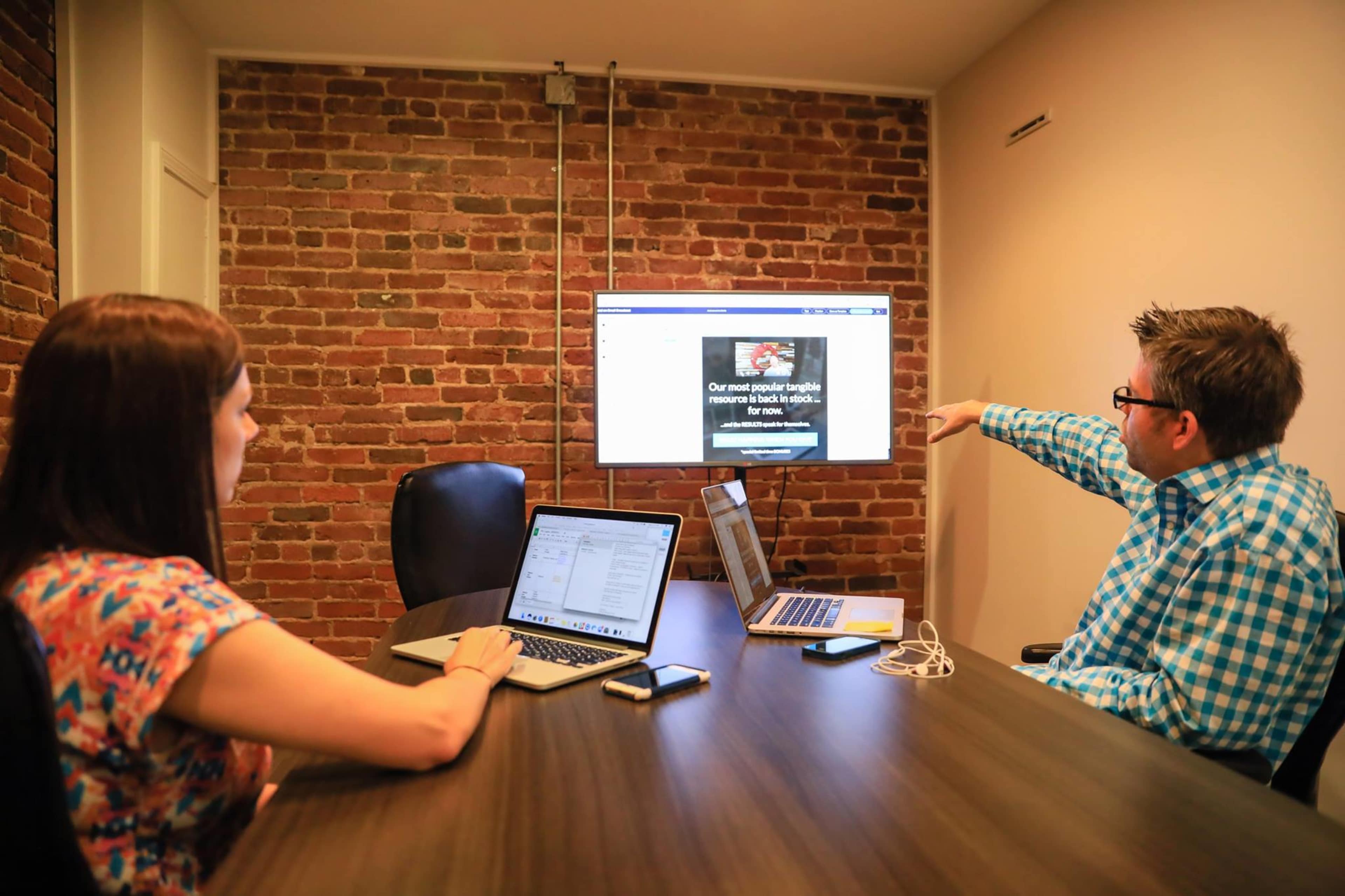 Two individuals sit at a conference table, with one pointing towards a screen displaying a website, while laptops are open in front of them.
