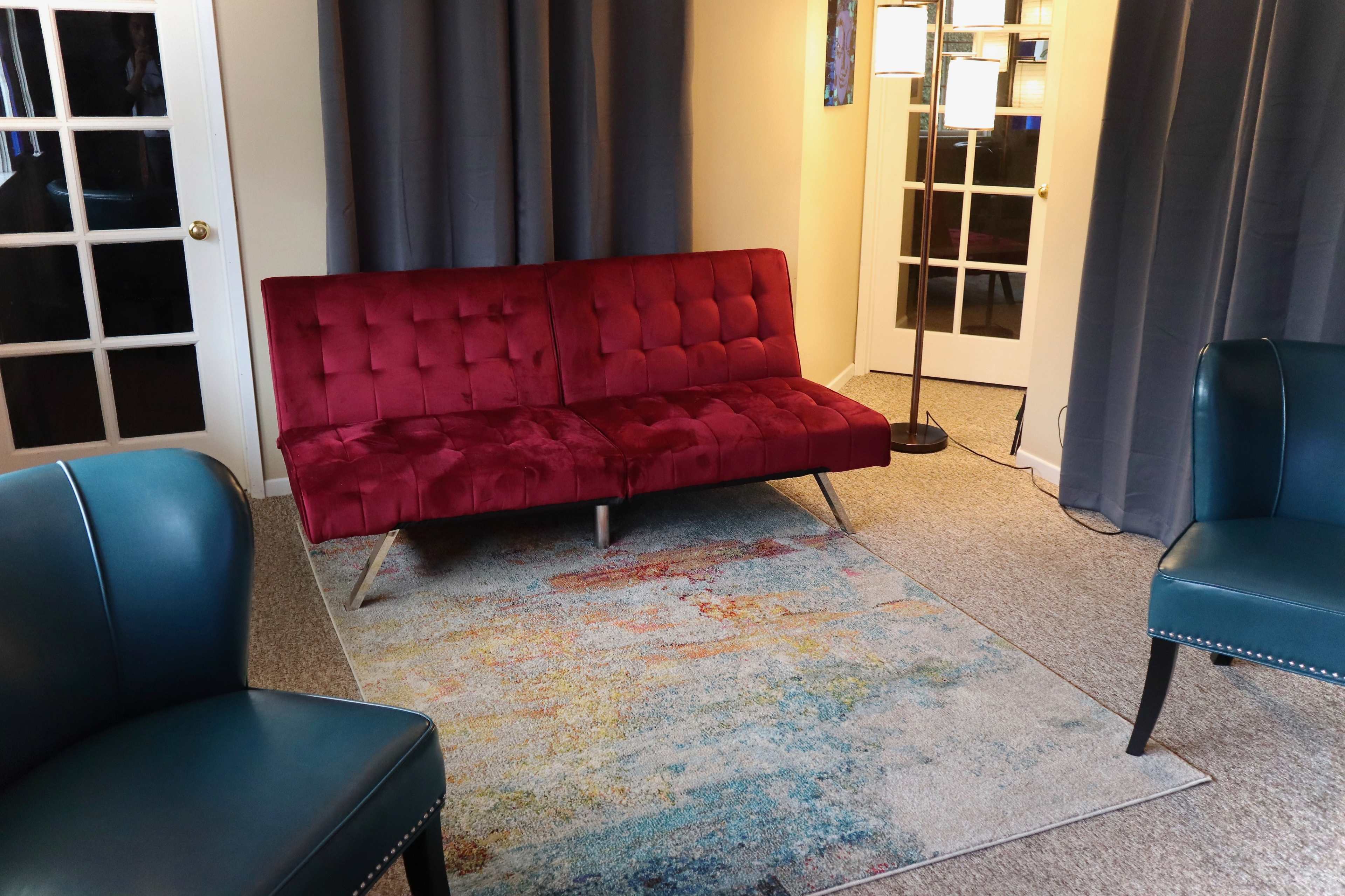 The image shows a living room with a red velvet couch positioned on a colorful area rug, accompanied by two teal armchairs and a standing lamp.