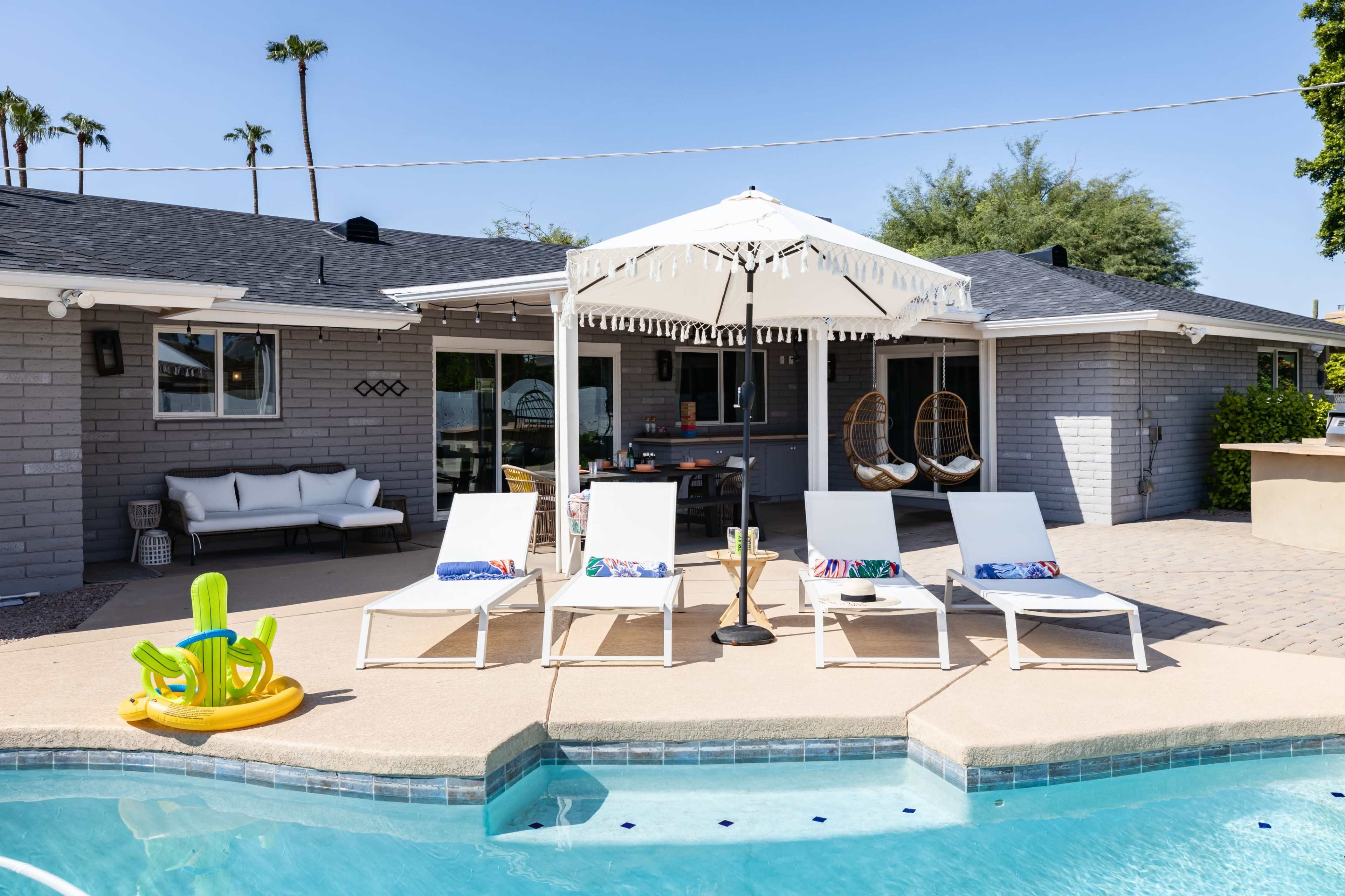 A swimming pool area features white lounge chairs and a decorative umbrella beside a modern house with a patio and palm trees in the background.