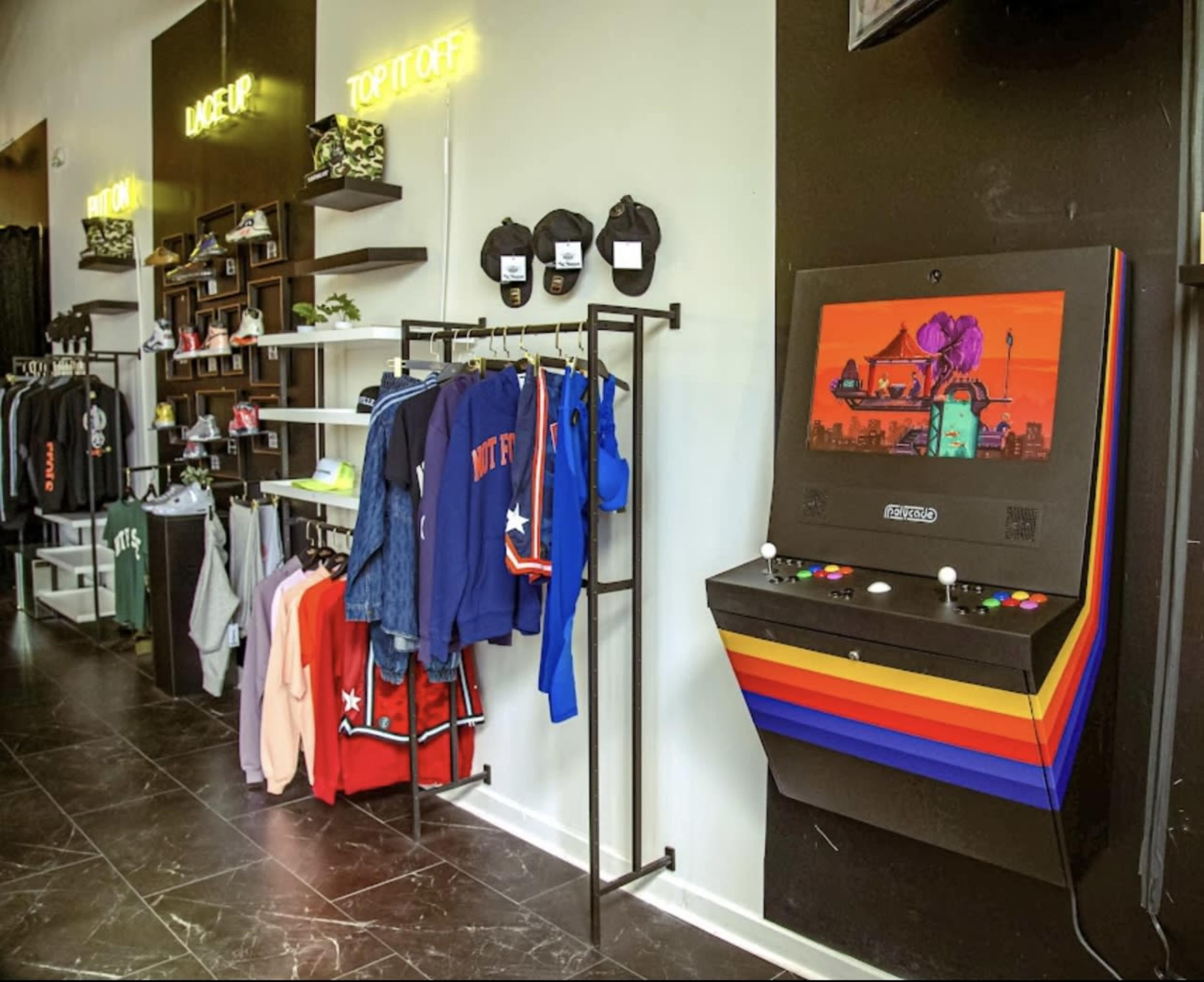 The image shows a modern retail space featuring clothing racks on the left and a vintage arcade game on the right.