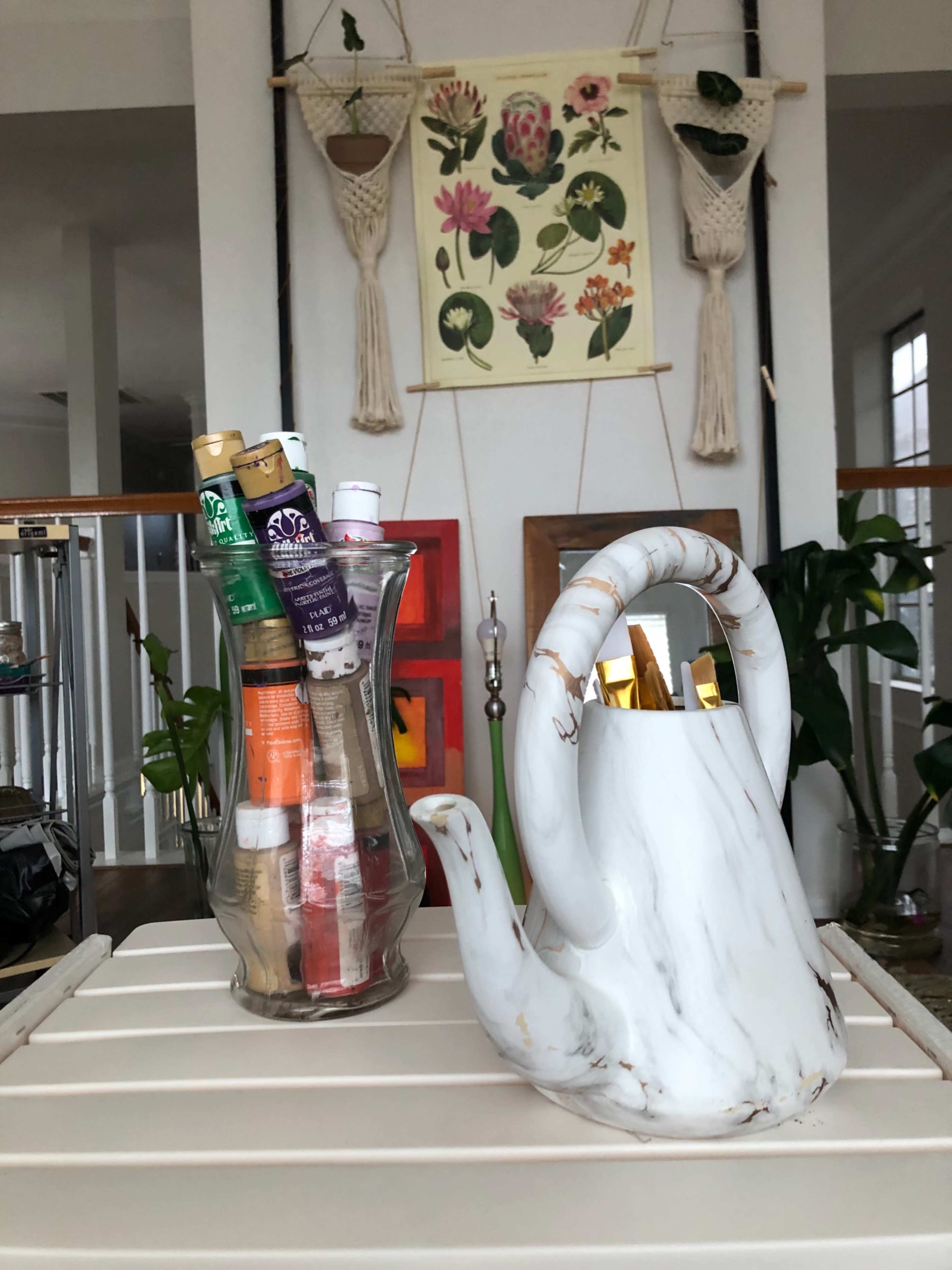 A marble-patterned watering can and a glass vase filled with rolled-up documents are displayed on a slatted table against a backdrop of plants and wall art.