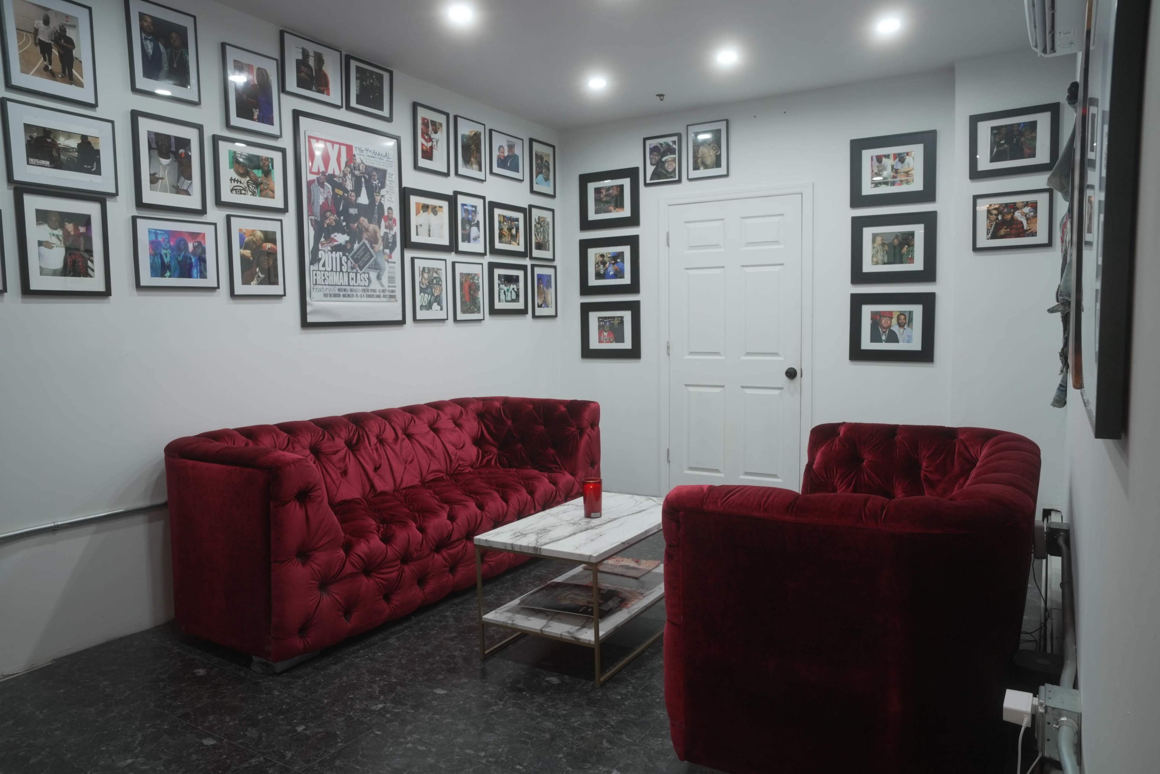 The room features two red velvet sofas positioned around a rectangular marble coffee table, with numerous framed photographs adorning the white walls.