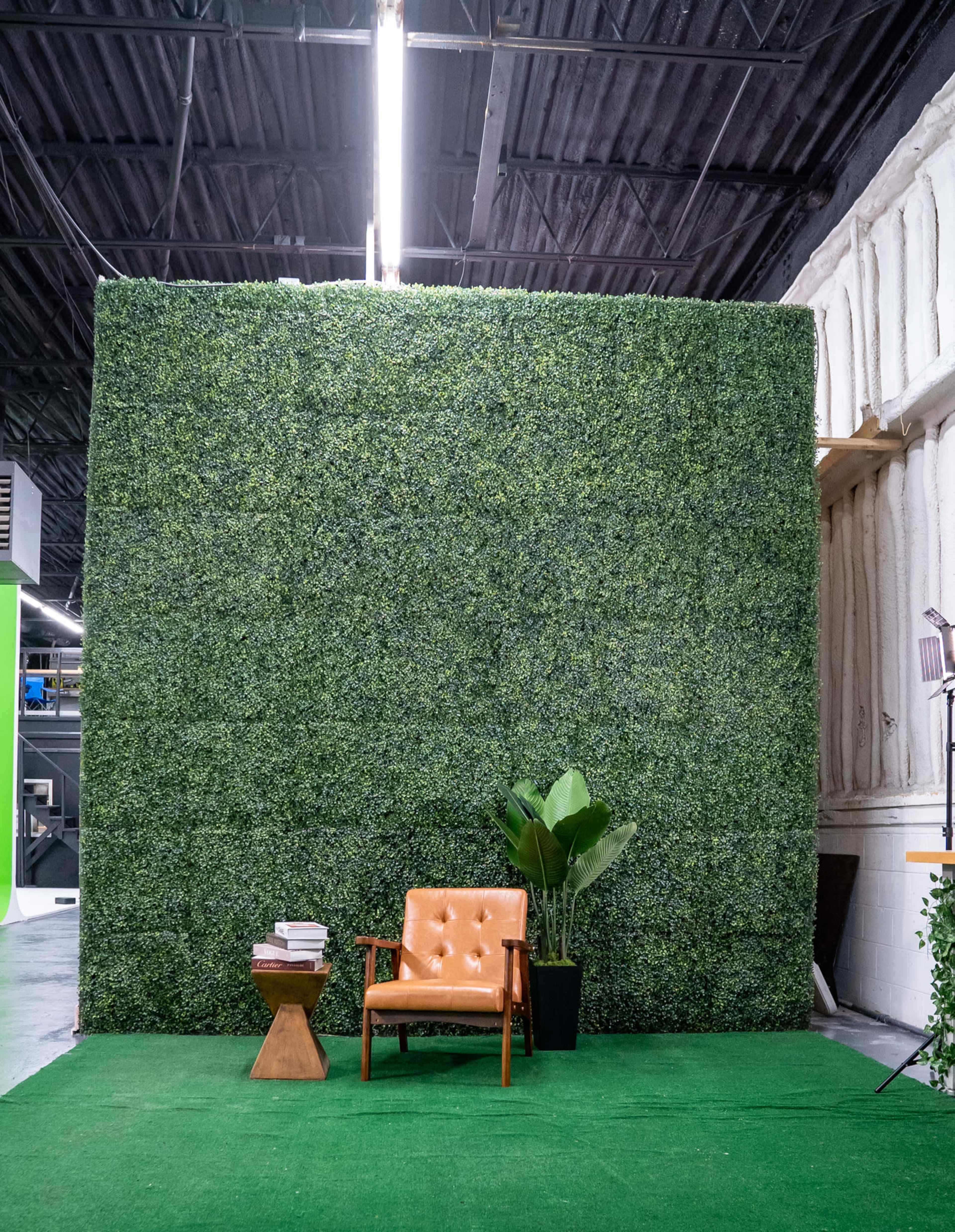 The image shows a lounge area featuring a brown armchair and a small wooden side table against a backdrop of a green hedge wall.