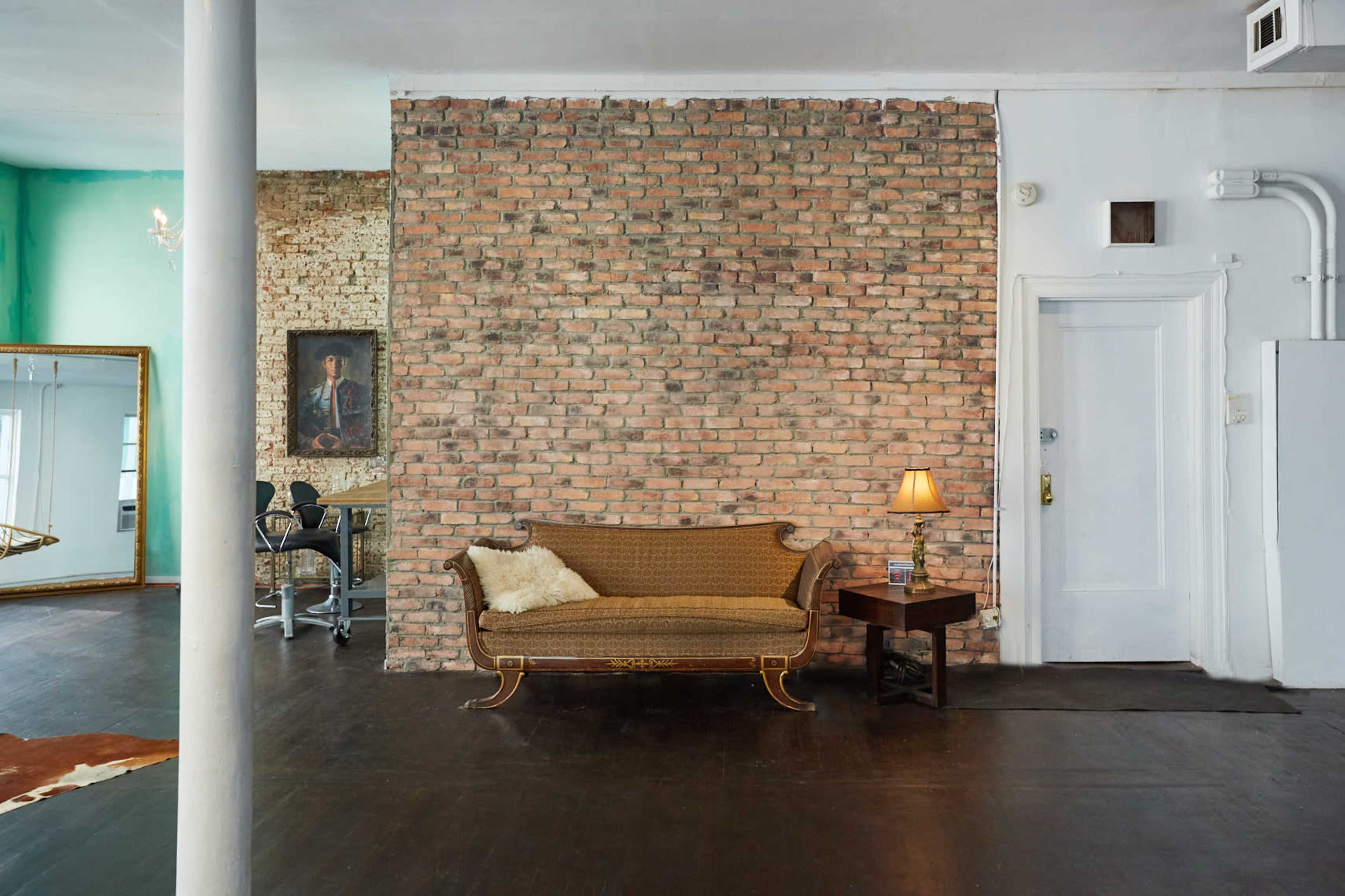 A stylish room with a brick wall, a vintage-style sofa, a side table with a lamp, and a mirror reflecting part of the space.