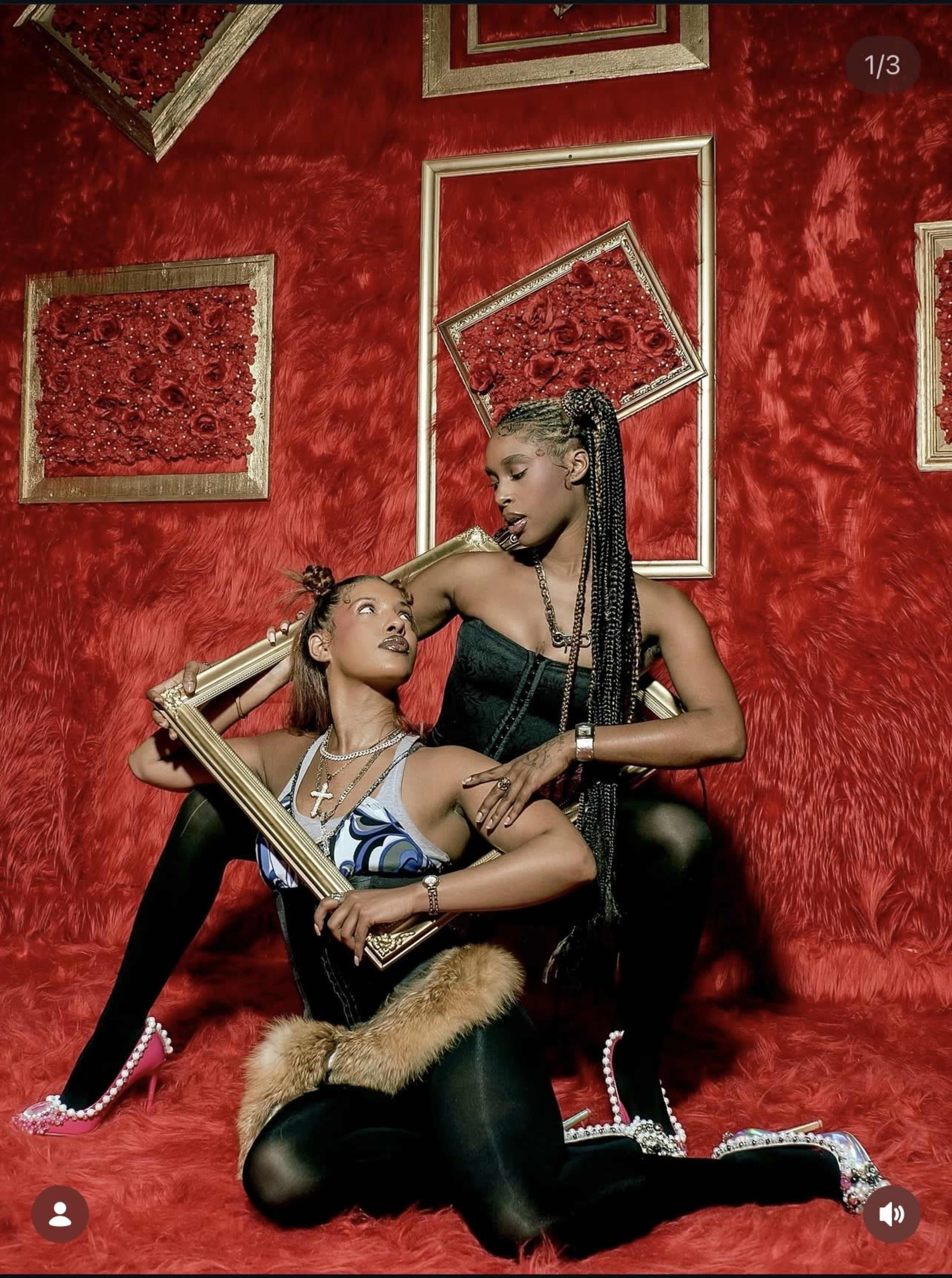 Two women are posed in front of a vibrant red textured backdrop, surrounded by ornate frames, one holding a decorative frame while the other leans into her pose.