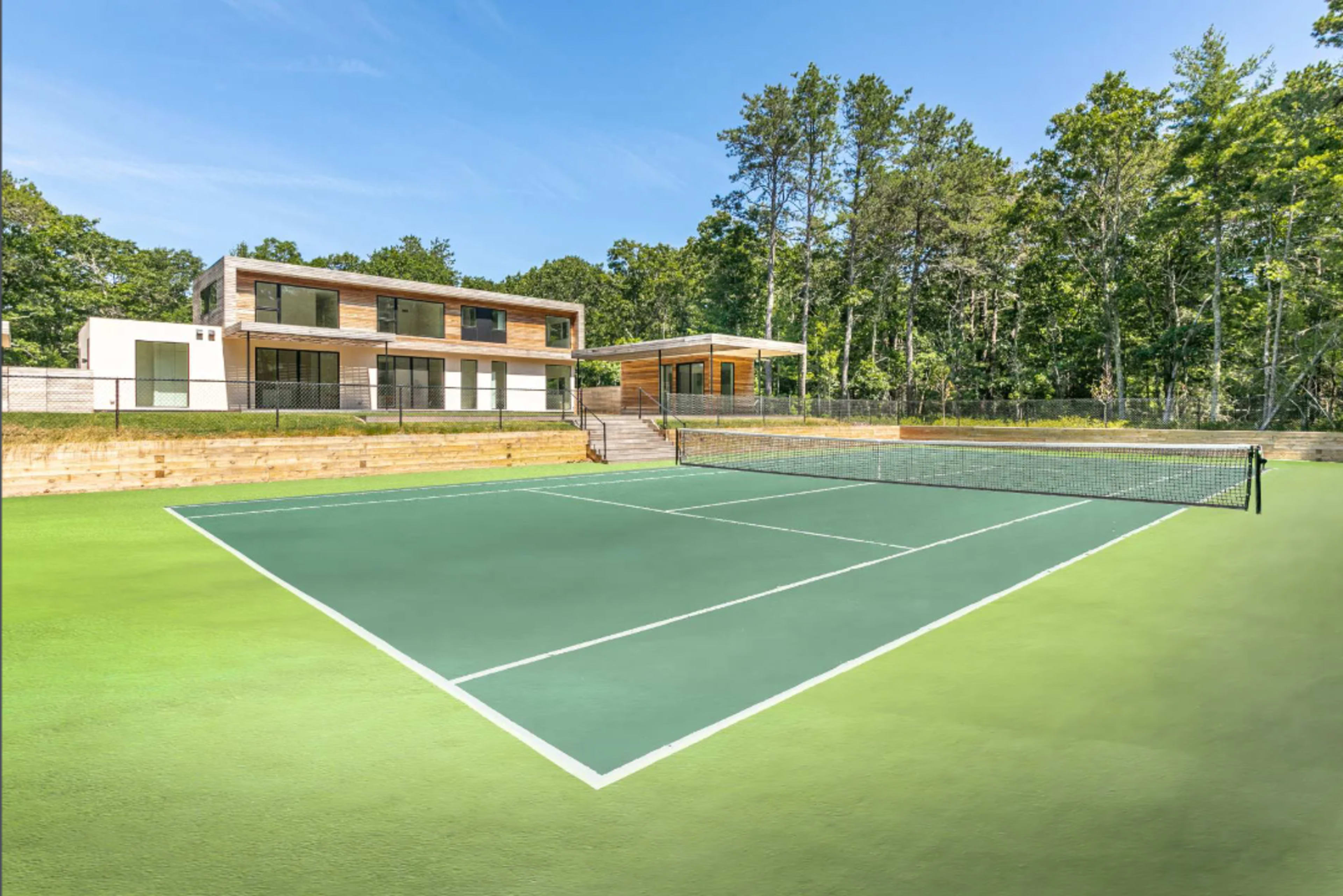A tennis court in front of a modern two-story house surrounded by trees.