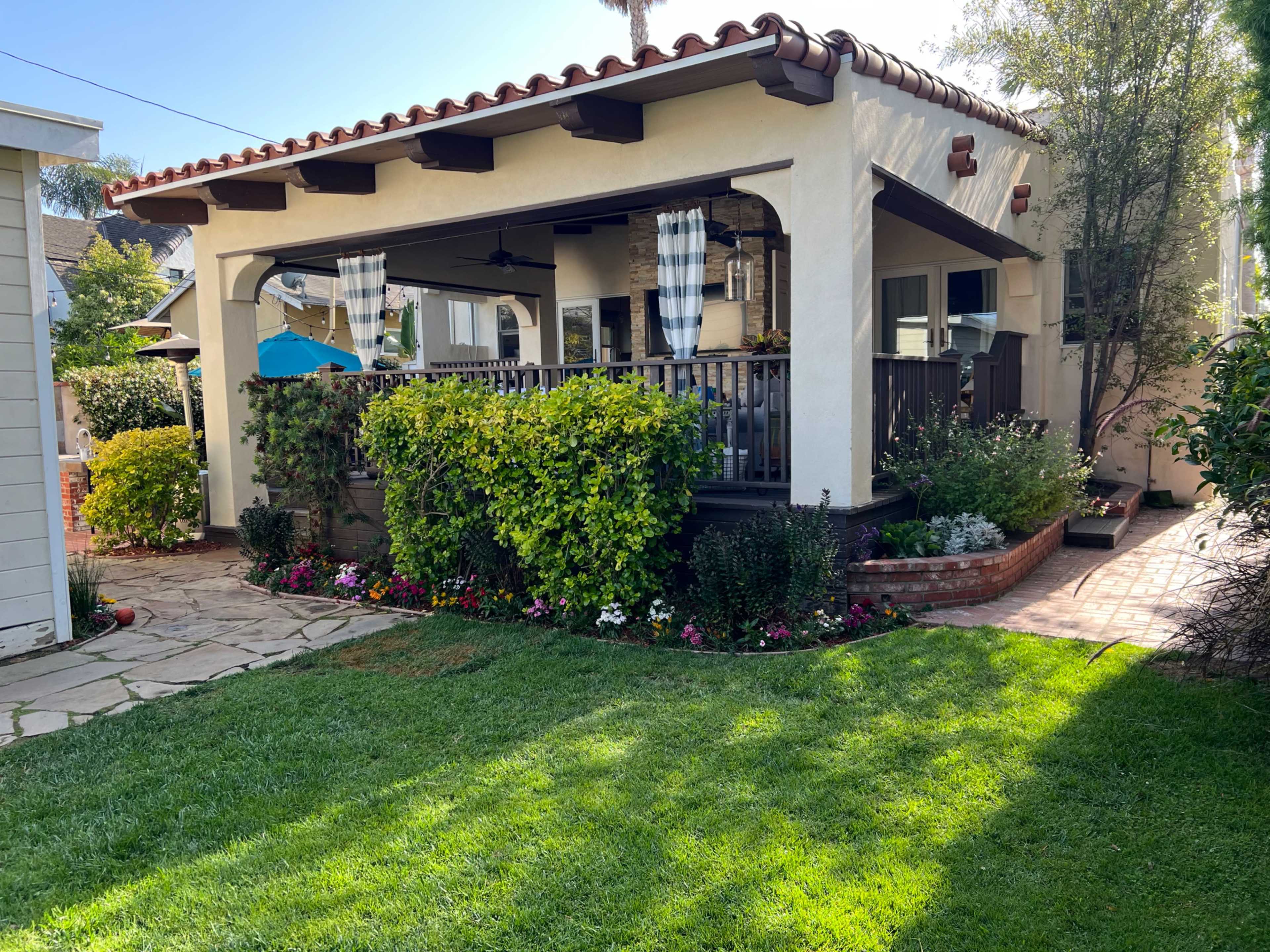 The image shows a well-maintained backyard featuring a patio with a covered seating area, surrounded by manicured grass and colorful flower beds.