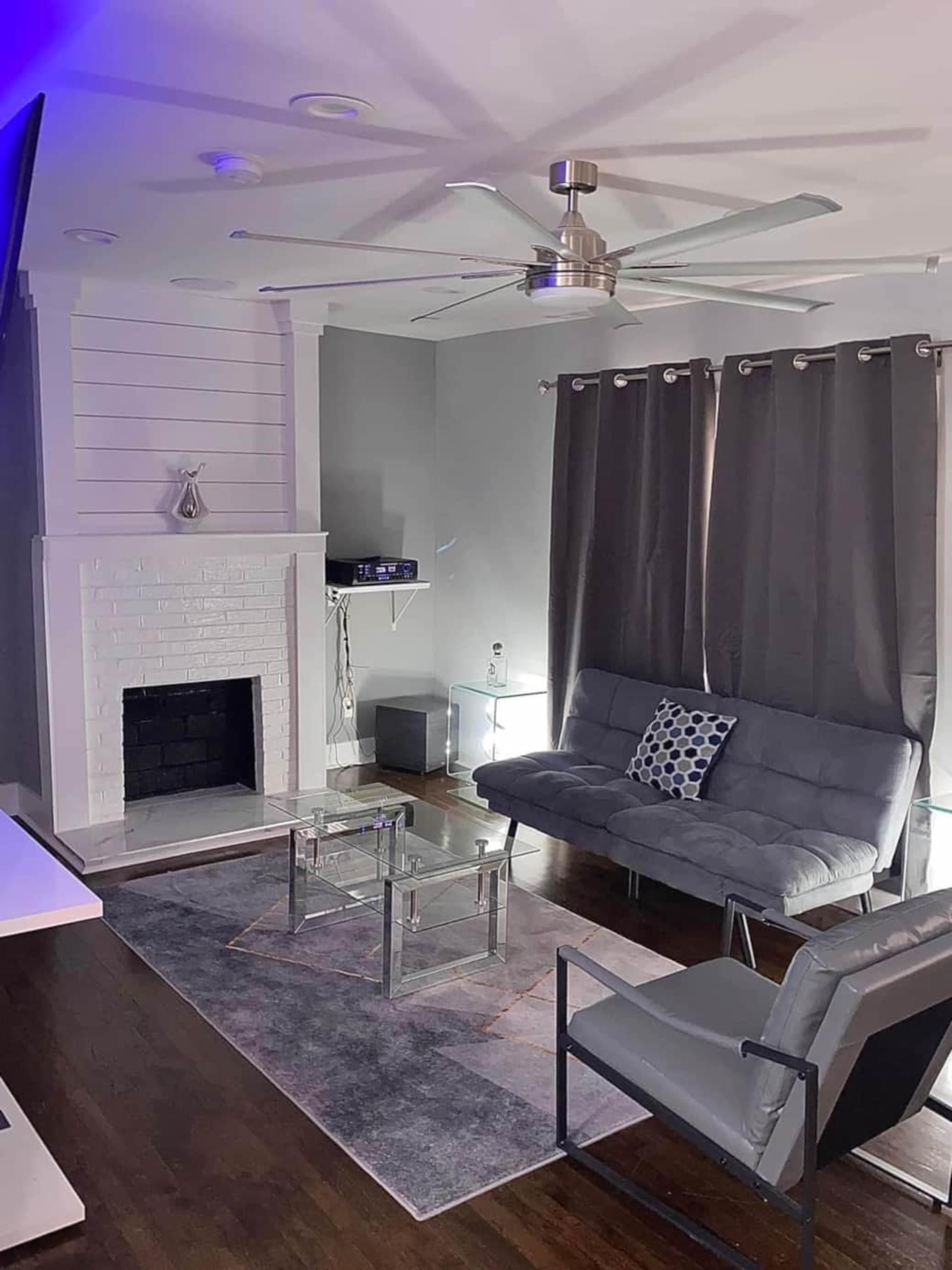 A modern living room featuring a gray sofa, a glass coffee table, a ceiling fan, and dark curtains, with a fireplace and wall-mounted shelves in the background.