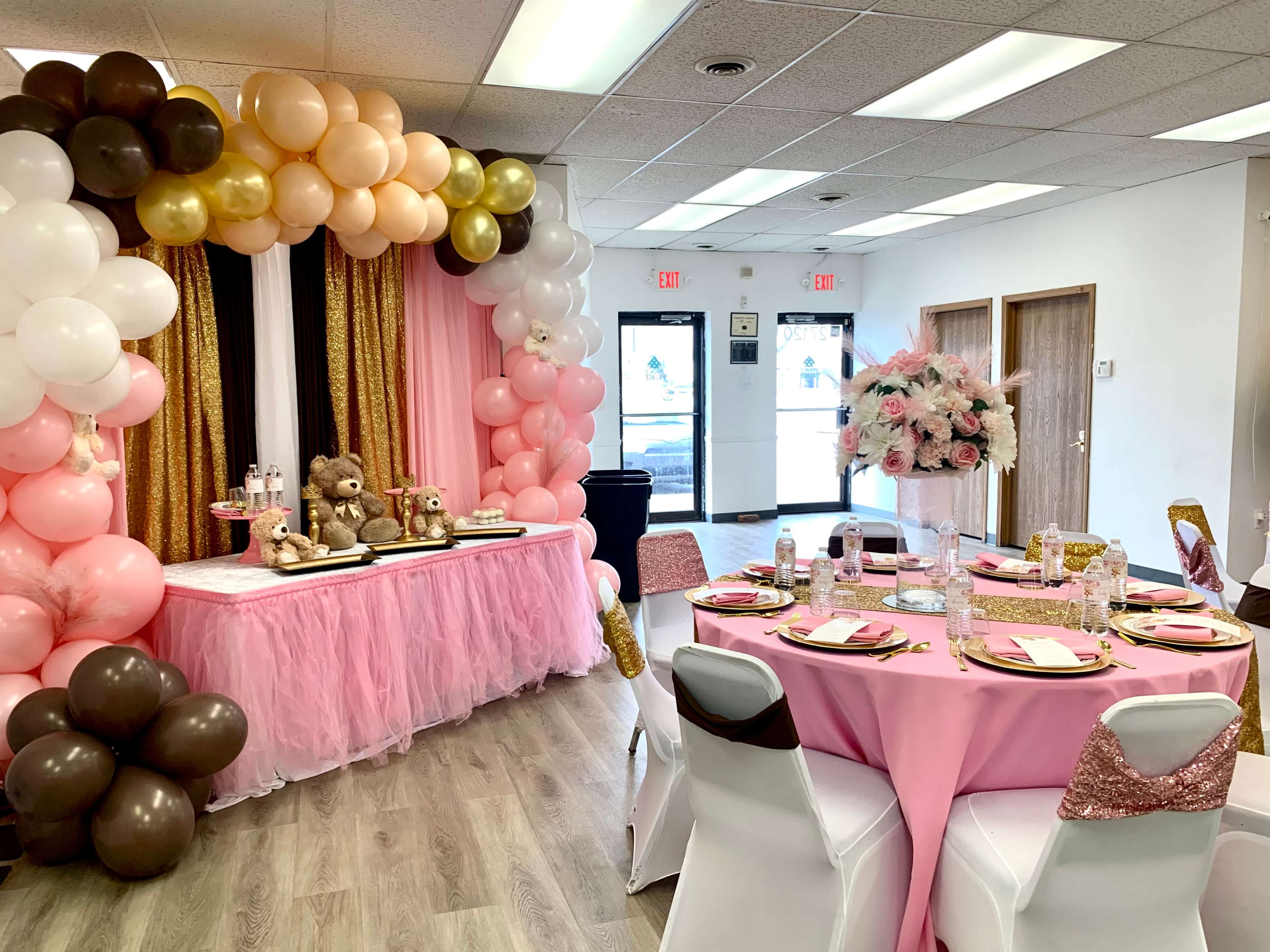 A decorated event space with pink and gold balloons, a table set for a celebration, and floral centerpieces.