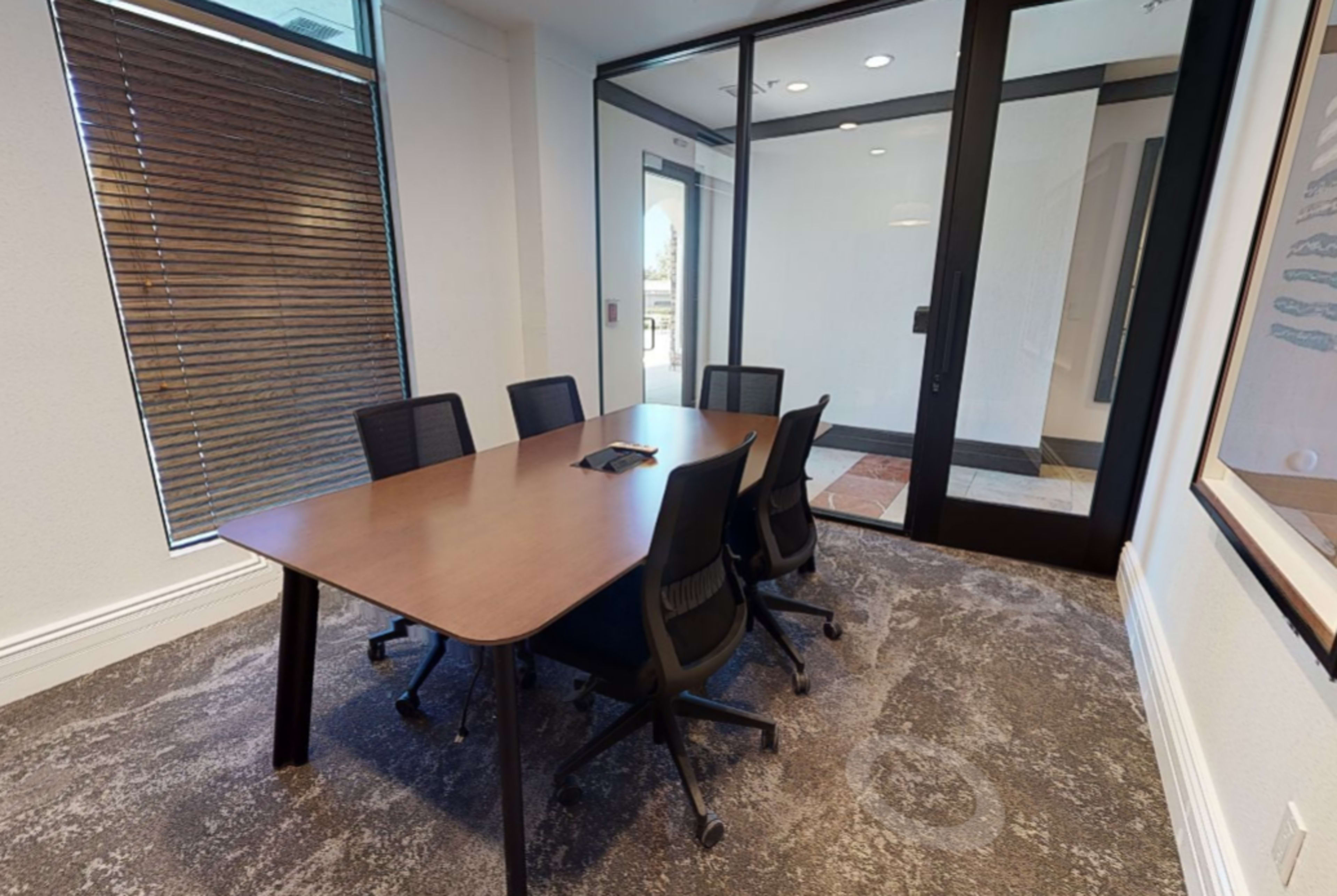 A conference room with a wooden table surrounded by six black chairs and a glass door leading to an exterior area.