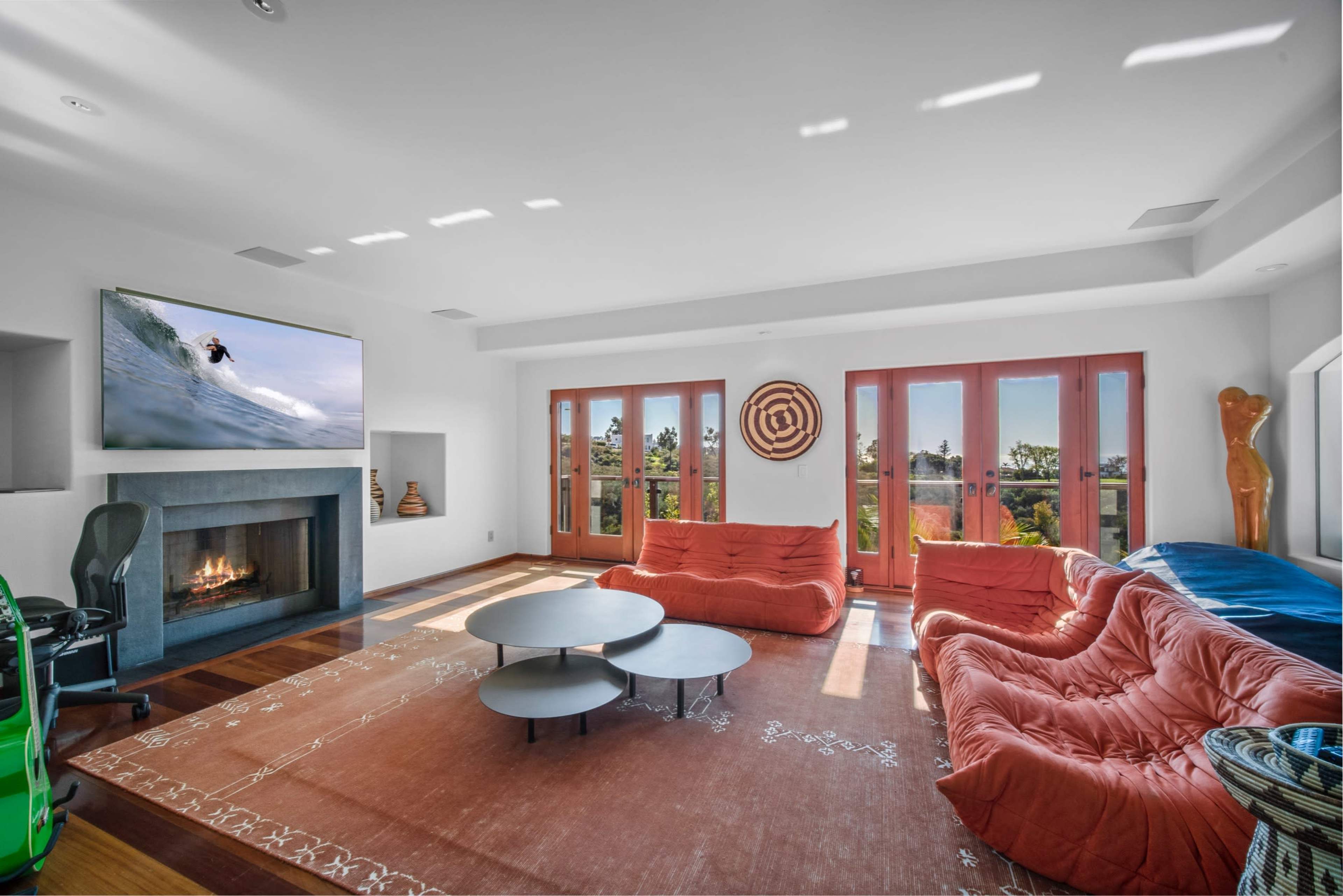 A modern living room features two orange sofas, a round coffee table, a fireplace, and large windows overlooking an outdoor view.