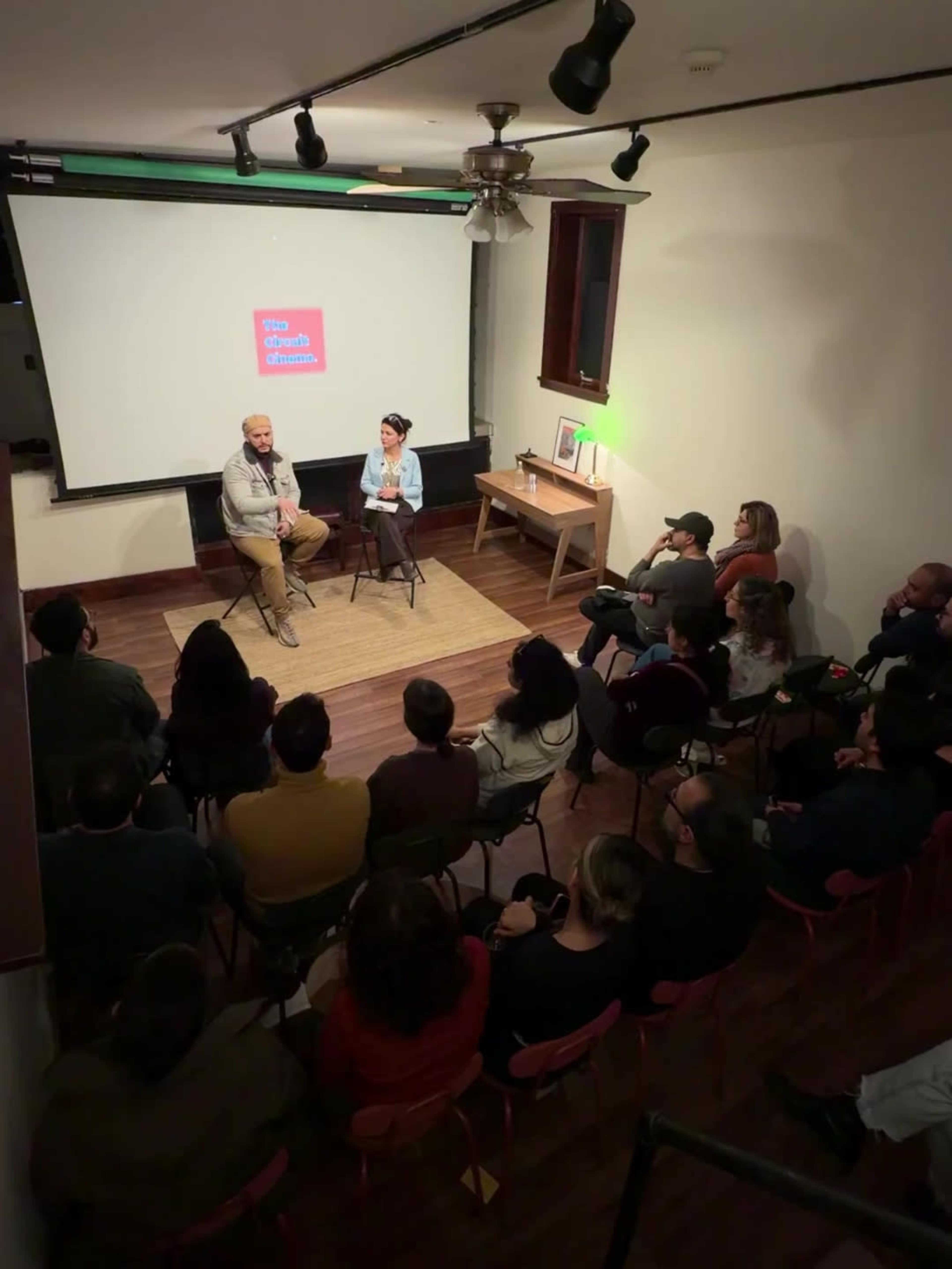 A speaker and a moderator sit on a stage in front of an audience during an event in a small, well-lit room.
