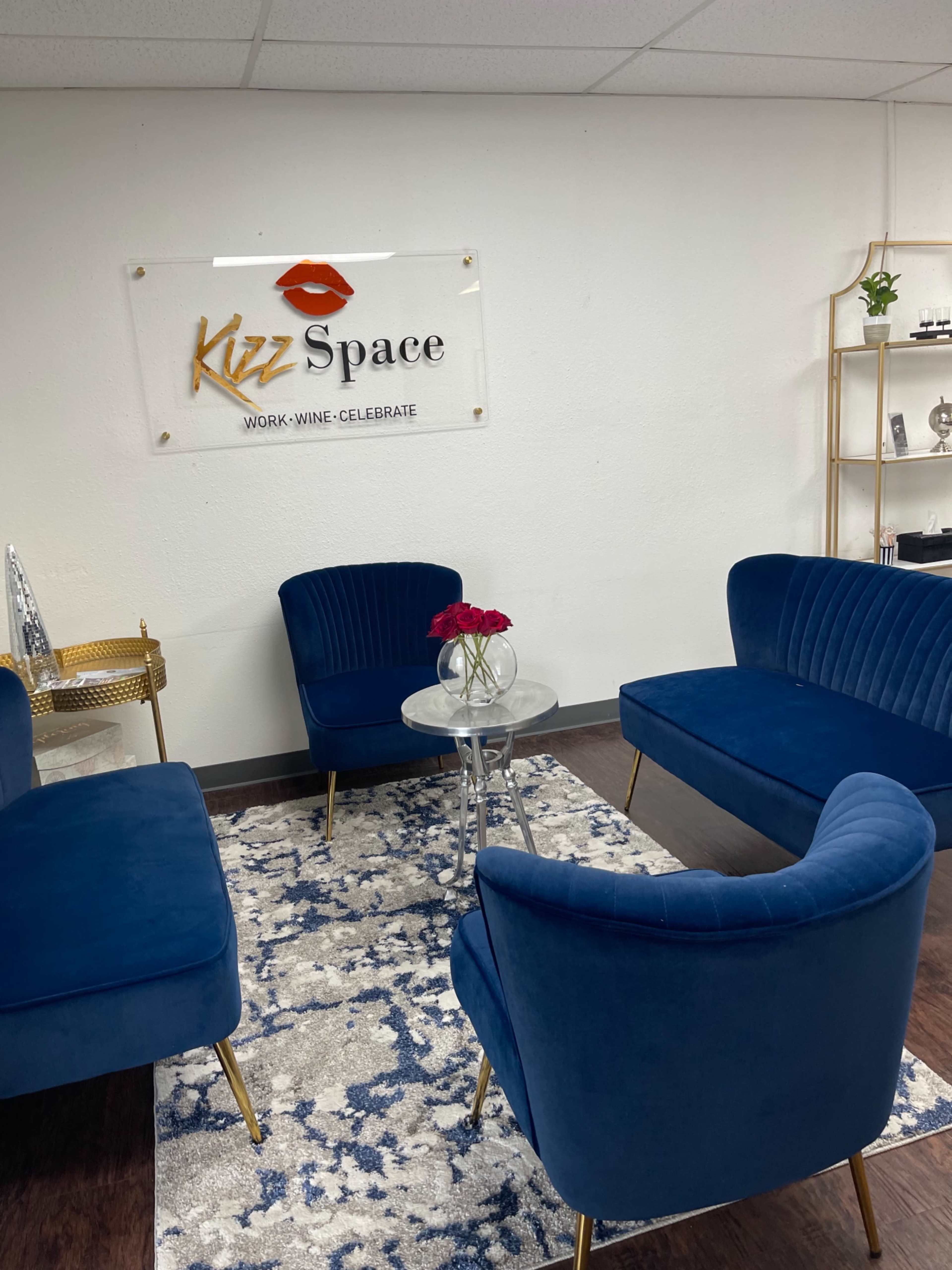 The image shows a modern lounge area with blue velvet chairs arranged around a glass table on a patterned rug, featuring a sign for "Kizz Space" on the wall.