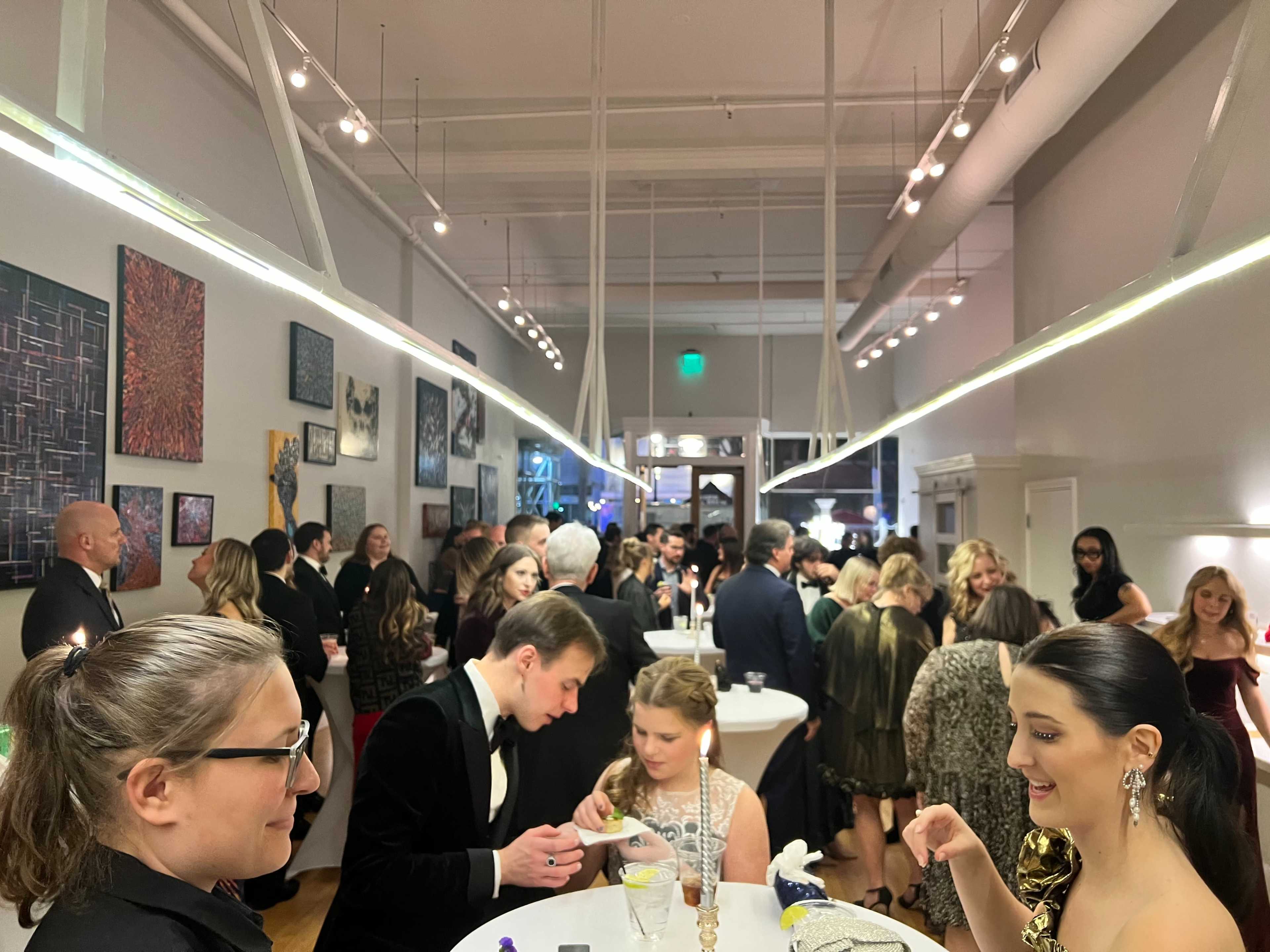 A crowd of elegantly dressed individuals mingles at a social event in an art gallery, surrounded by colorful artworks on the walls.