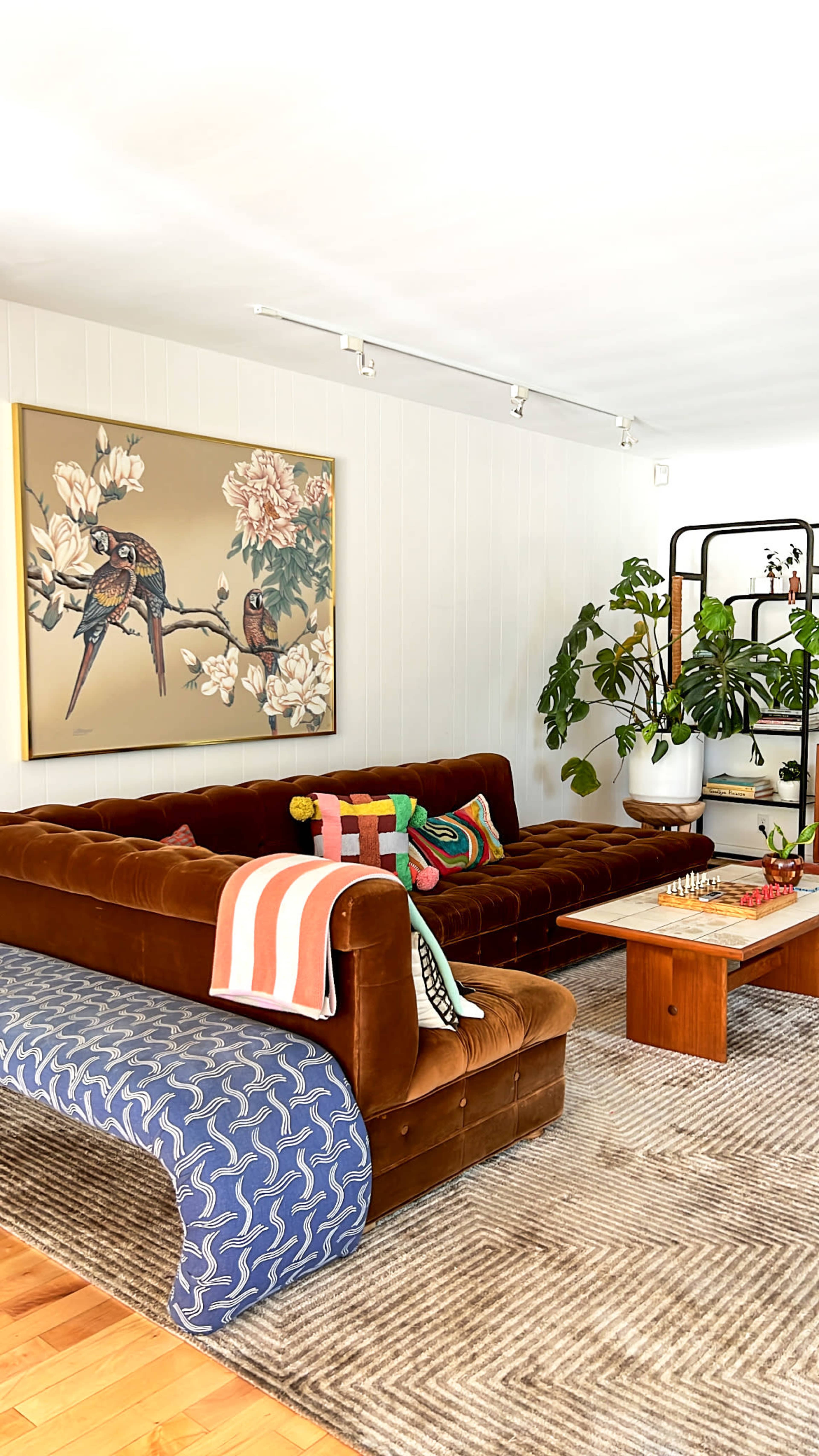 A cozy living room features a brown velvet sofa adorned with colorful pillows, a nature-themed artwork on the wall, and a plant beside a wooden coffee table on a patterned rug.