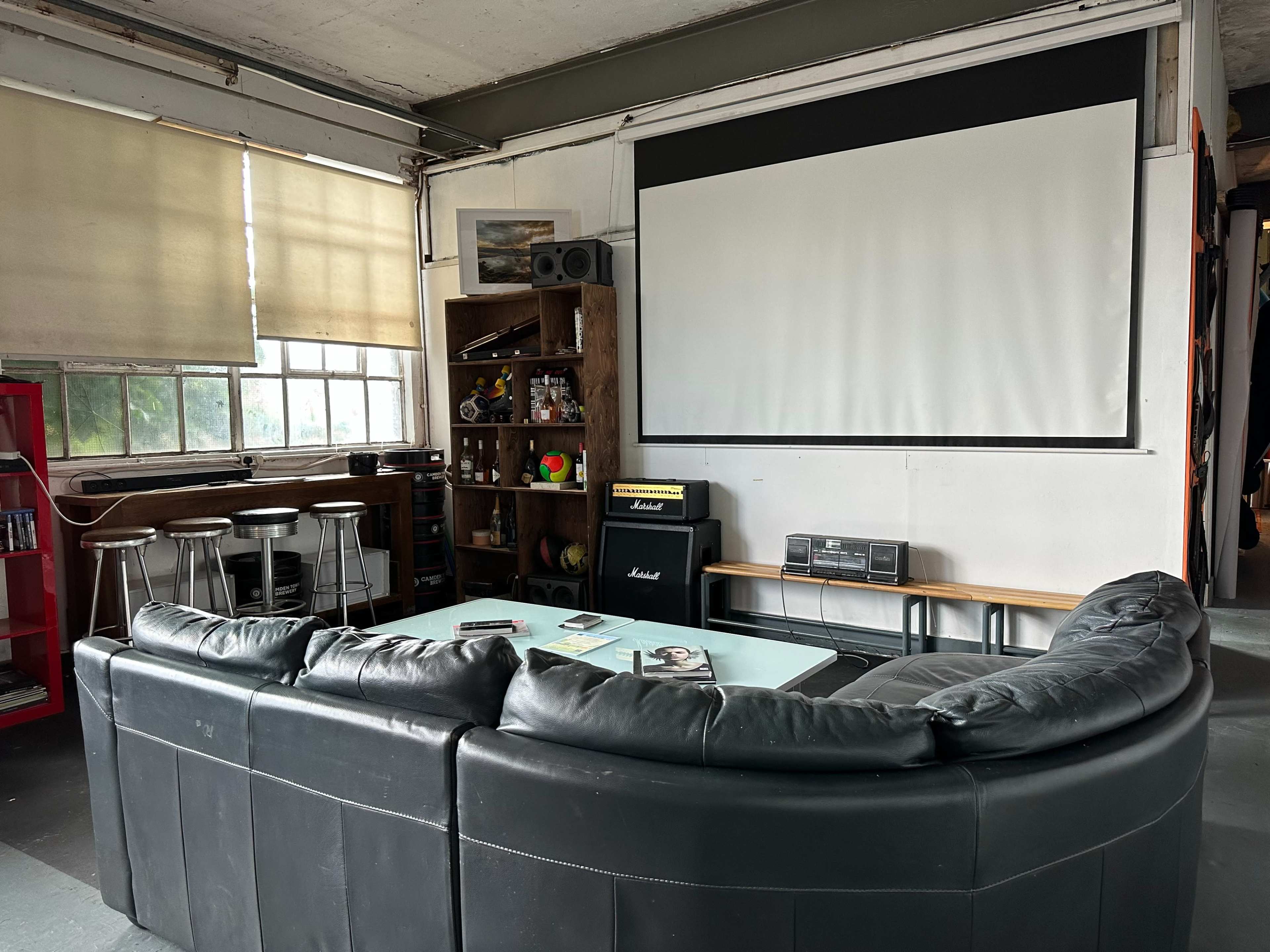 A spacious room with a black leather sectional couch facing a large blank projection screen, accompanied by a wooden shelf filled with various items and a bar area with stools in the background.