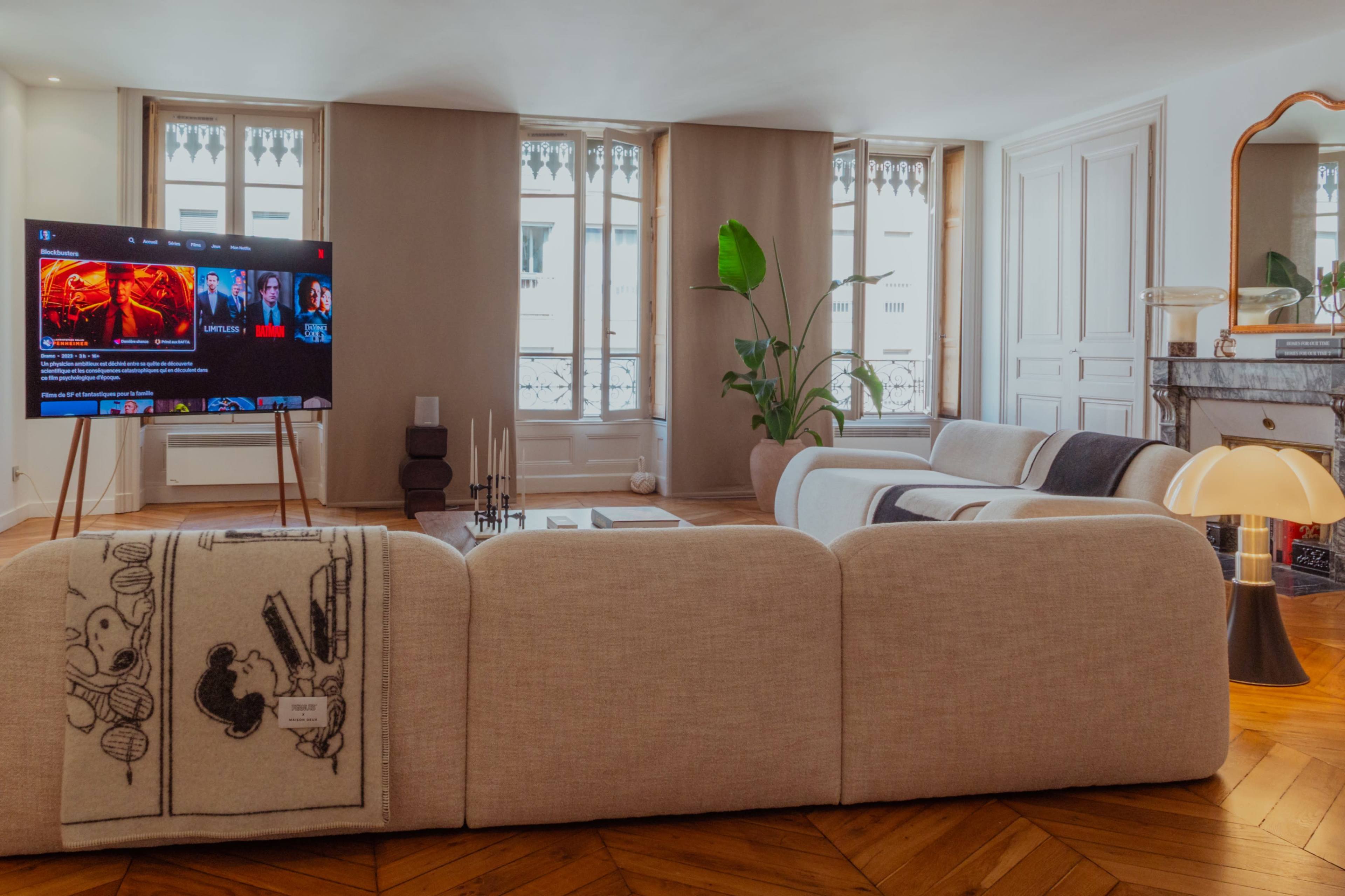 A modern living room features a large sectional sofa, a television displaying streaming options, and a decorative mirror, with large windows allowing natural light to enter the space.