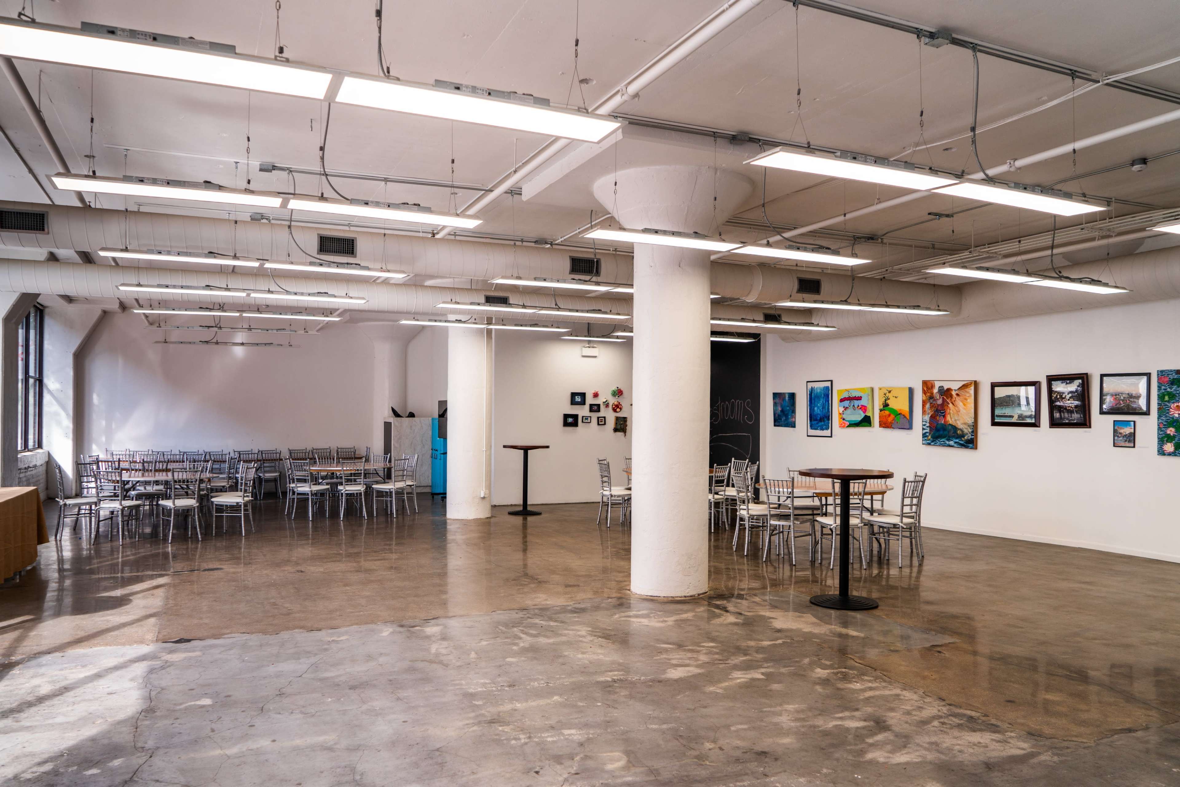 The image shows a spacious, airy gallery or event space with tables and chairs arranged in two distinct areas, and walls adorned with various artworks.