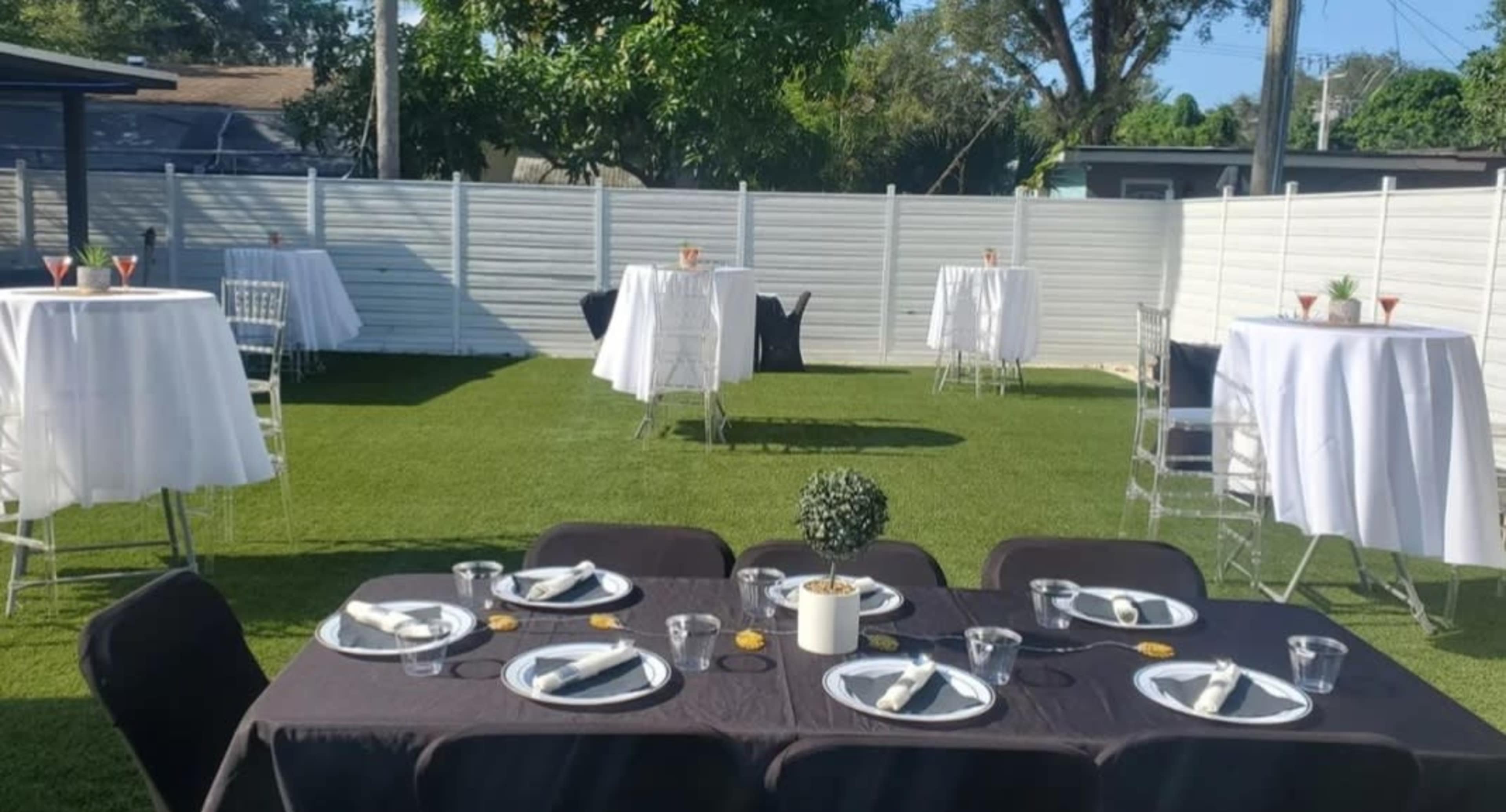The image depicts a backyard event setup featuring several decorated tables and chairs arranged on a lawn.