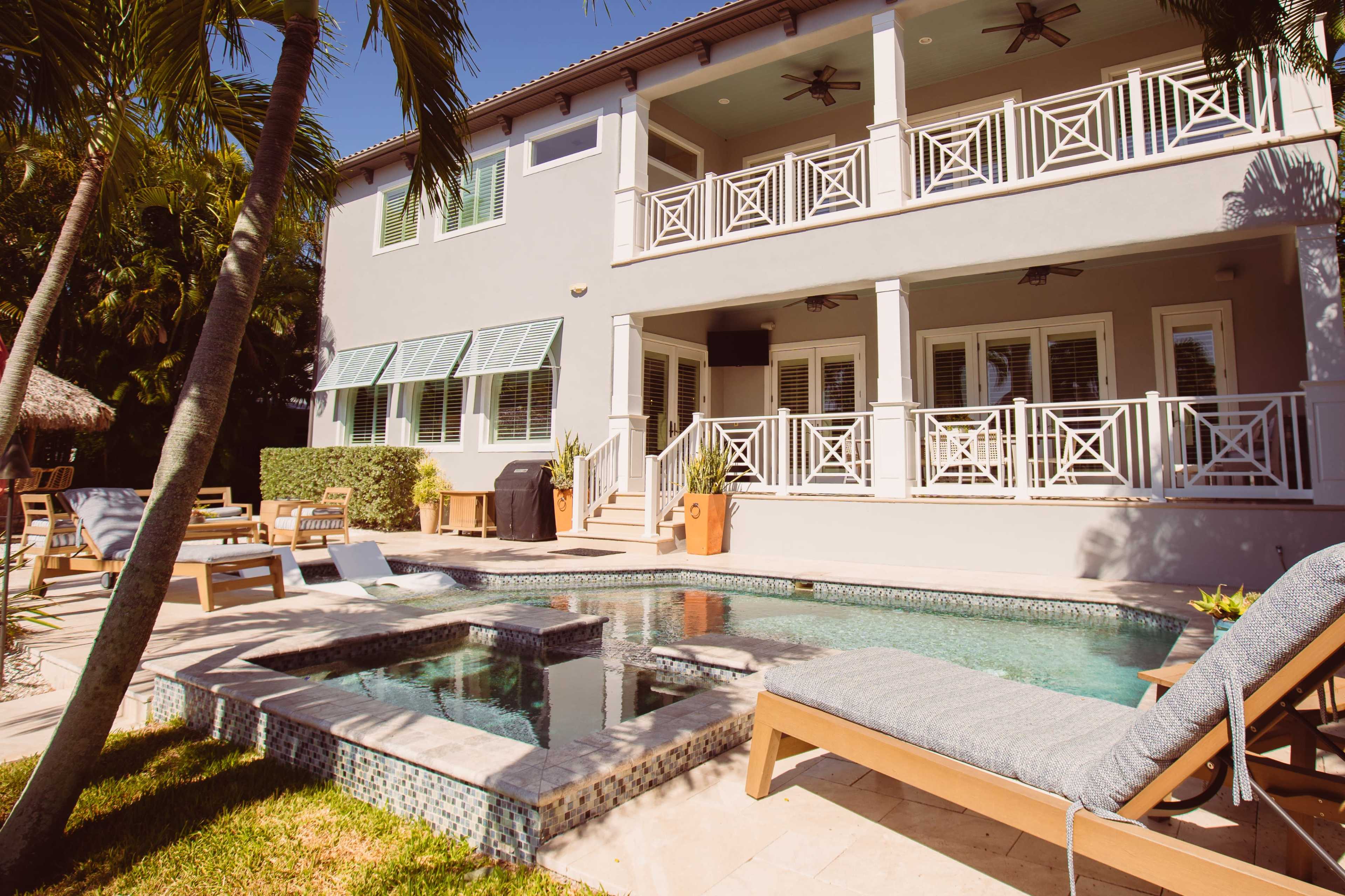 The image shows a modern house with a pool, surrounded by palm trees and lounge chairs, featuring a covered patio area.