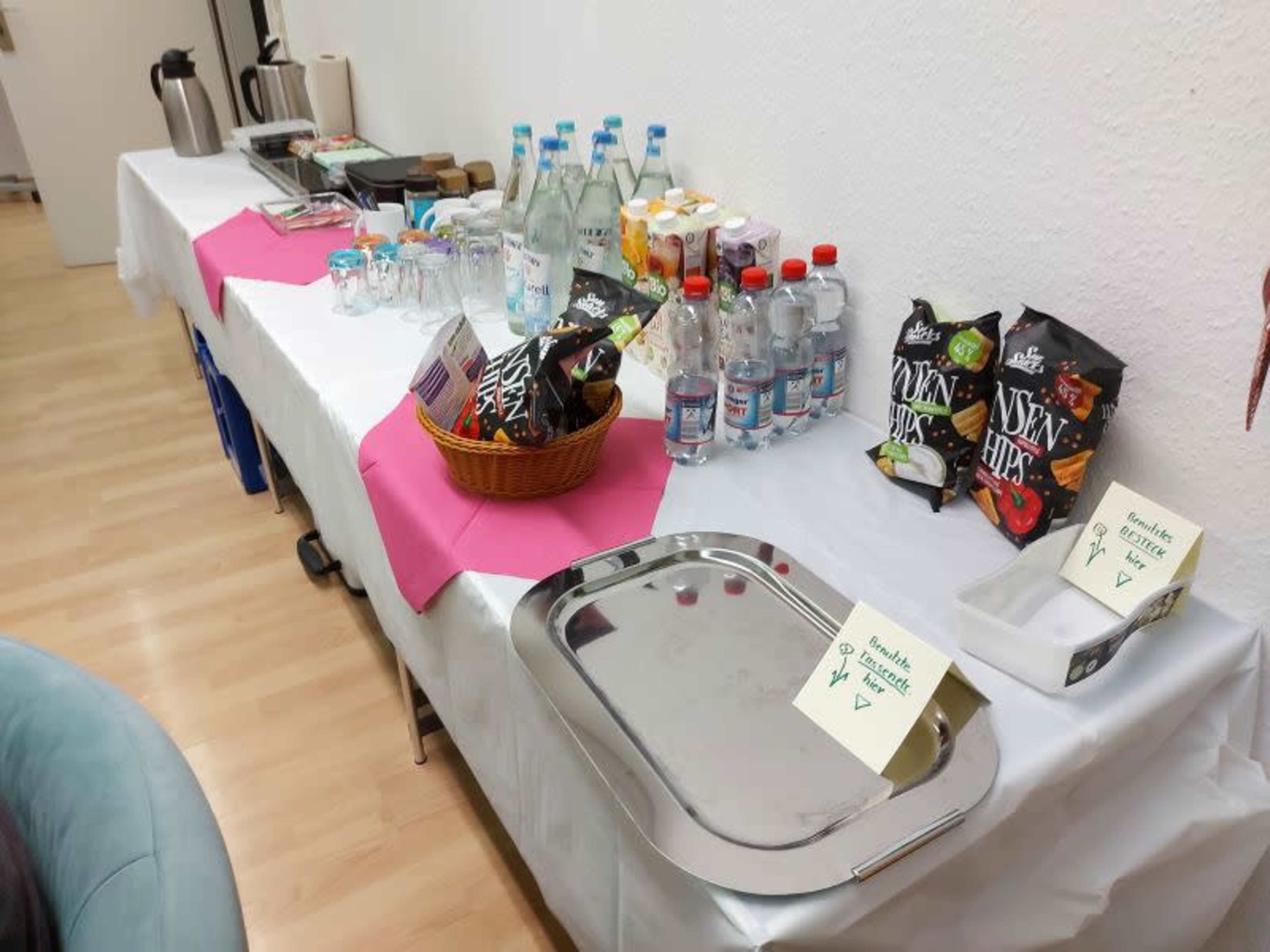 A table is set with snacks, drinks, and utensils for an event, featuring various bottles of water, snacks in bags, and several glasses.