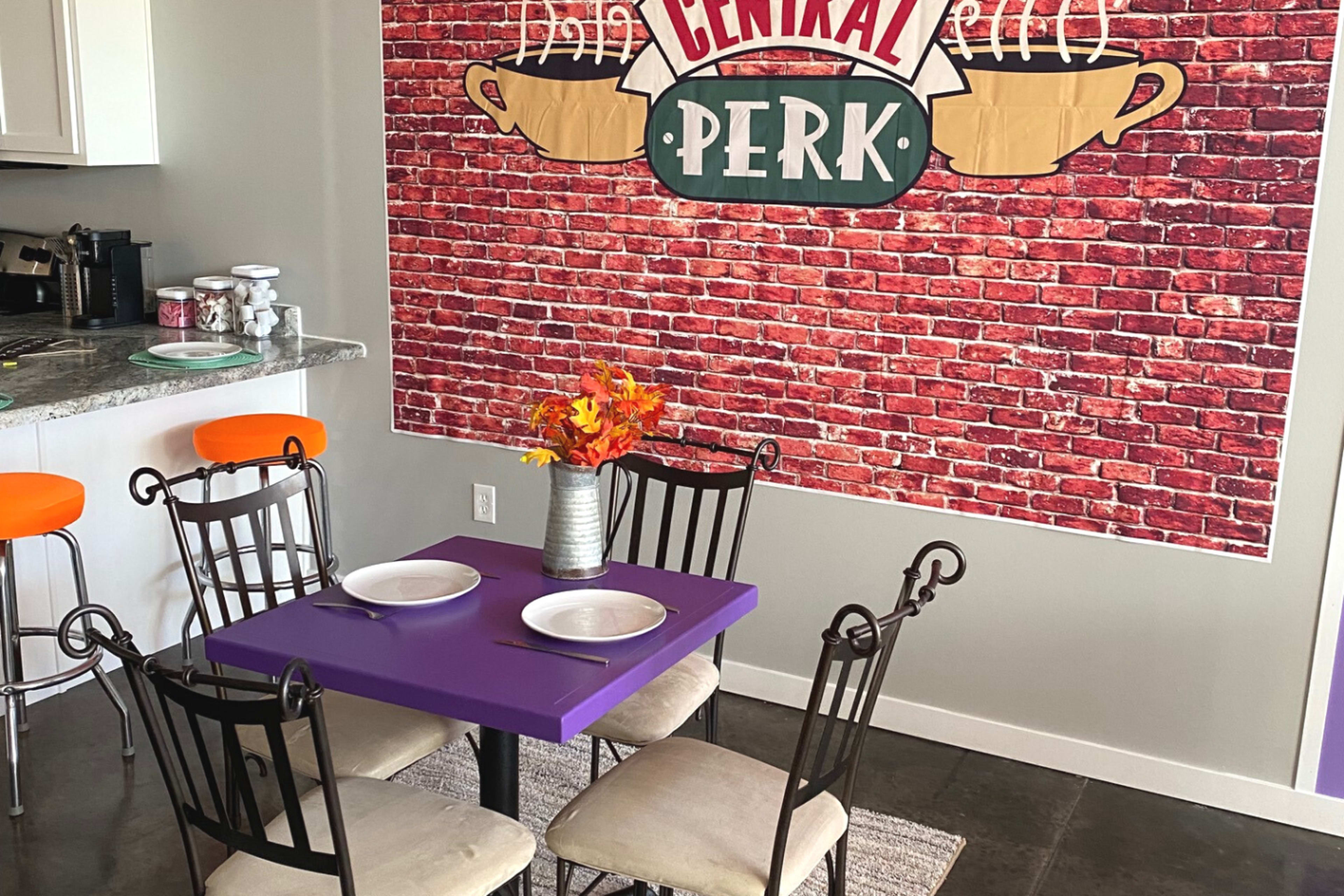 A table set with plates is positioned in front of a brick wall featuring a "Central Perk" coffee shop mural, with a kitchen counter and orange stools visible in the background.
