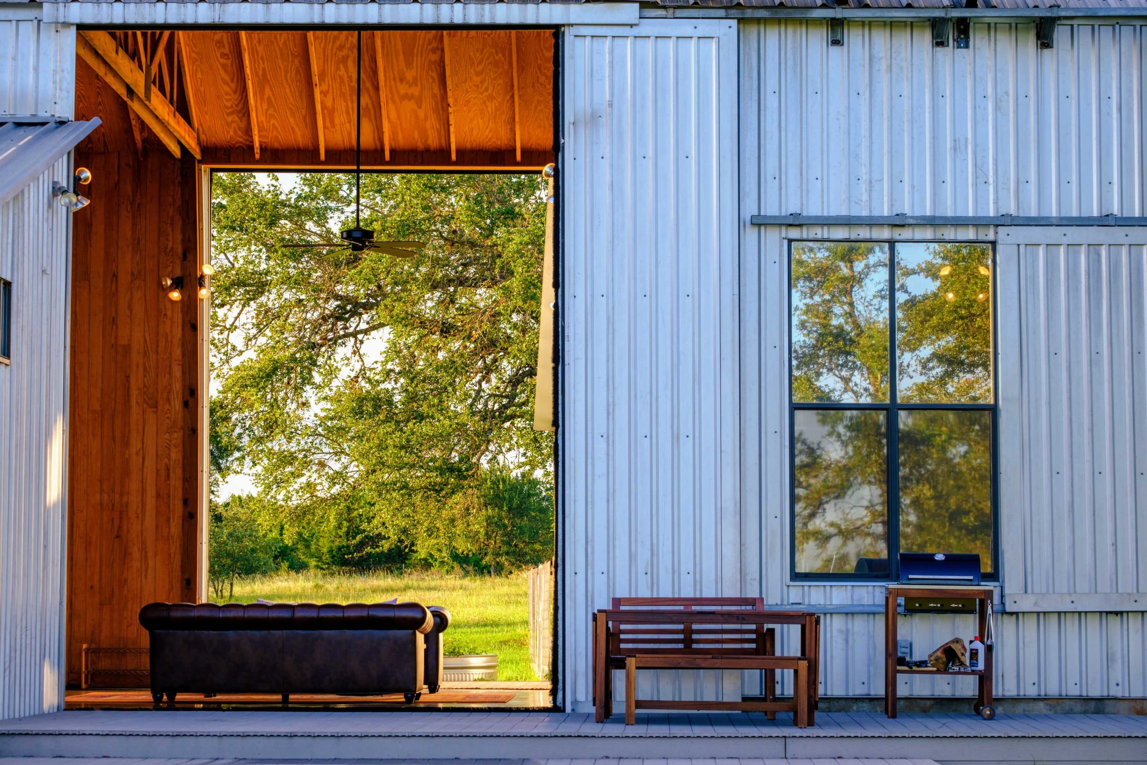 A cozy porch area featuring a leather sofa and a wooden bench beside a large opening that frames a view of a lush green field.