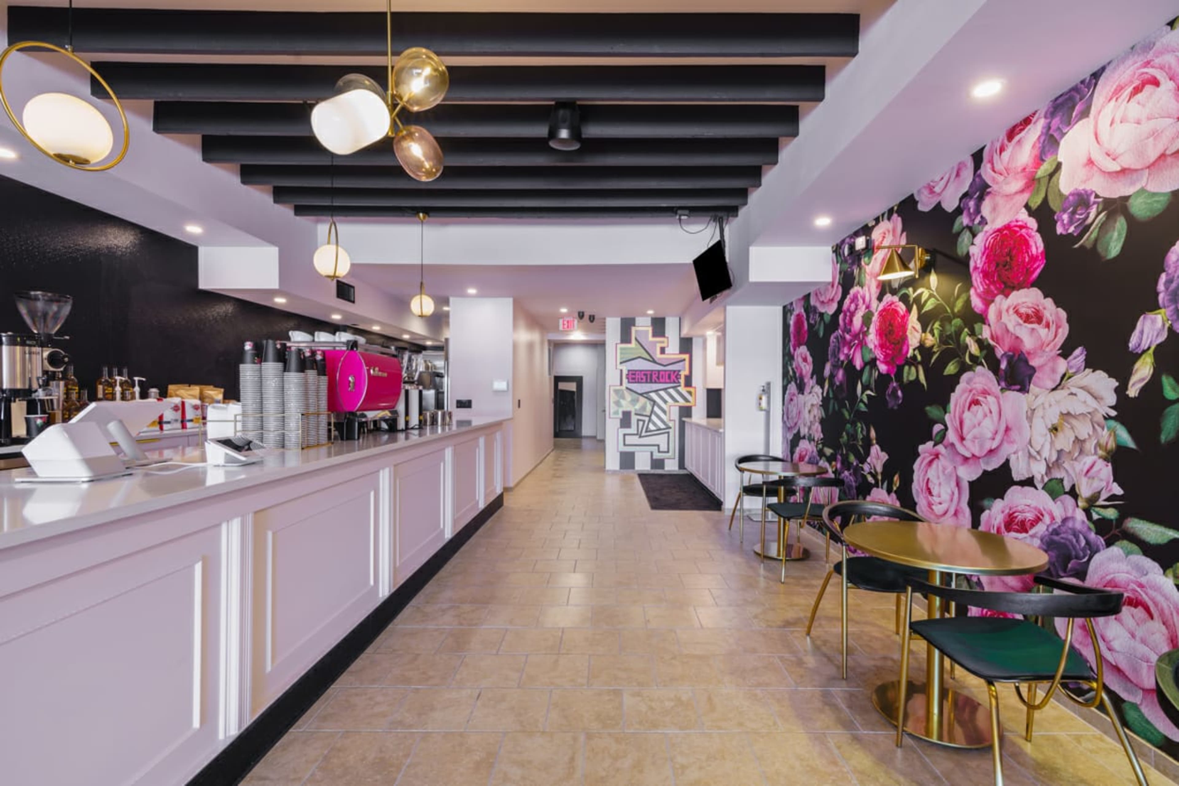 A modern café featuring a floral accent wall, a long counter with coffee equipment, and a seating area with round tables.