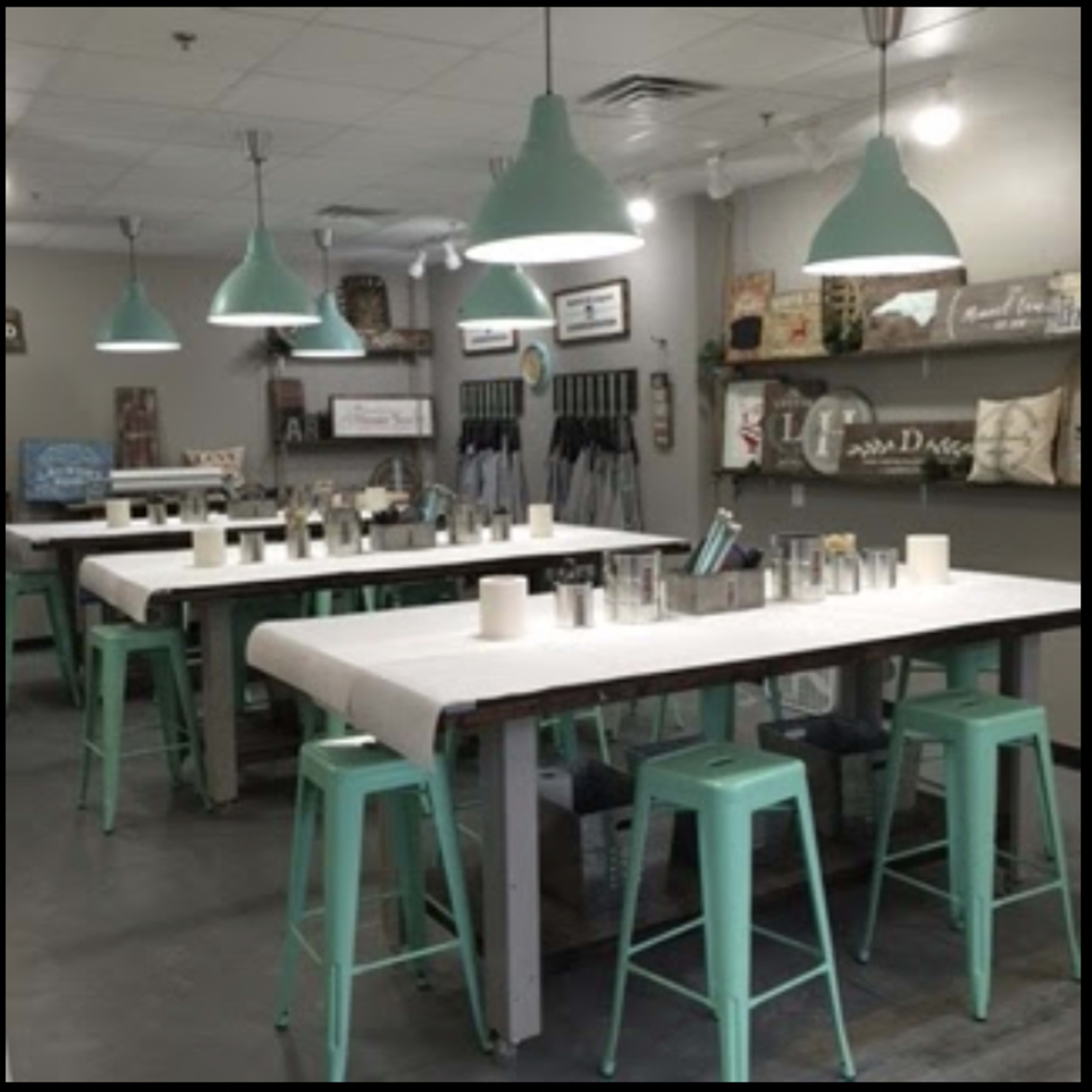 A spacious room with several large tables, mint-colored stools, and pendant lights hanging from the ceiling, arranged for a casual dining or crafting event.