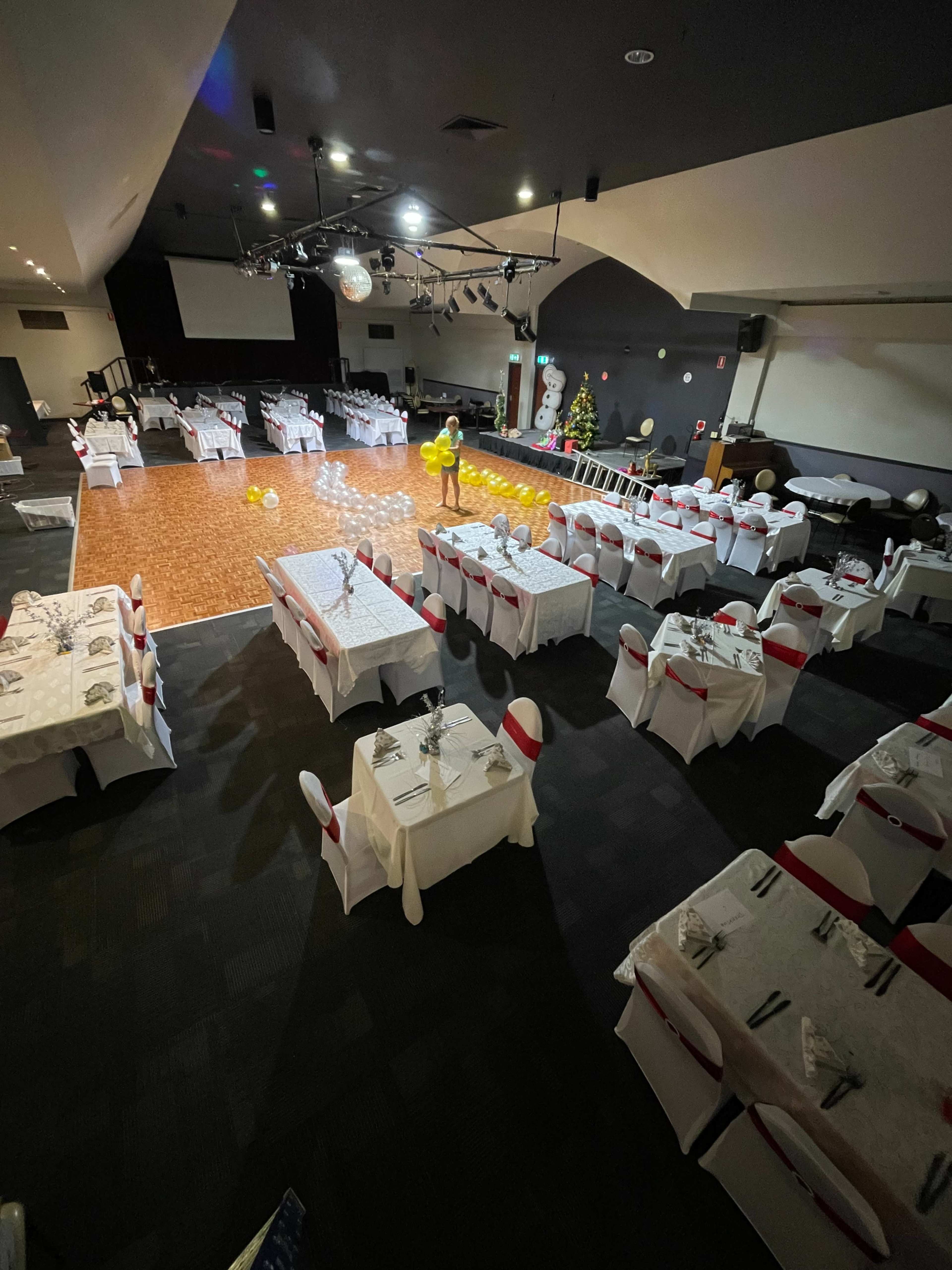 The image shows a banquet hall set up for an event, featuring tables with white tablecloths and red accents, arranged around a hardwood dance floor.