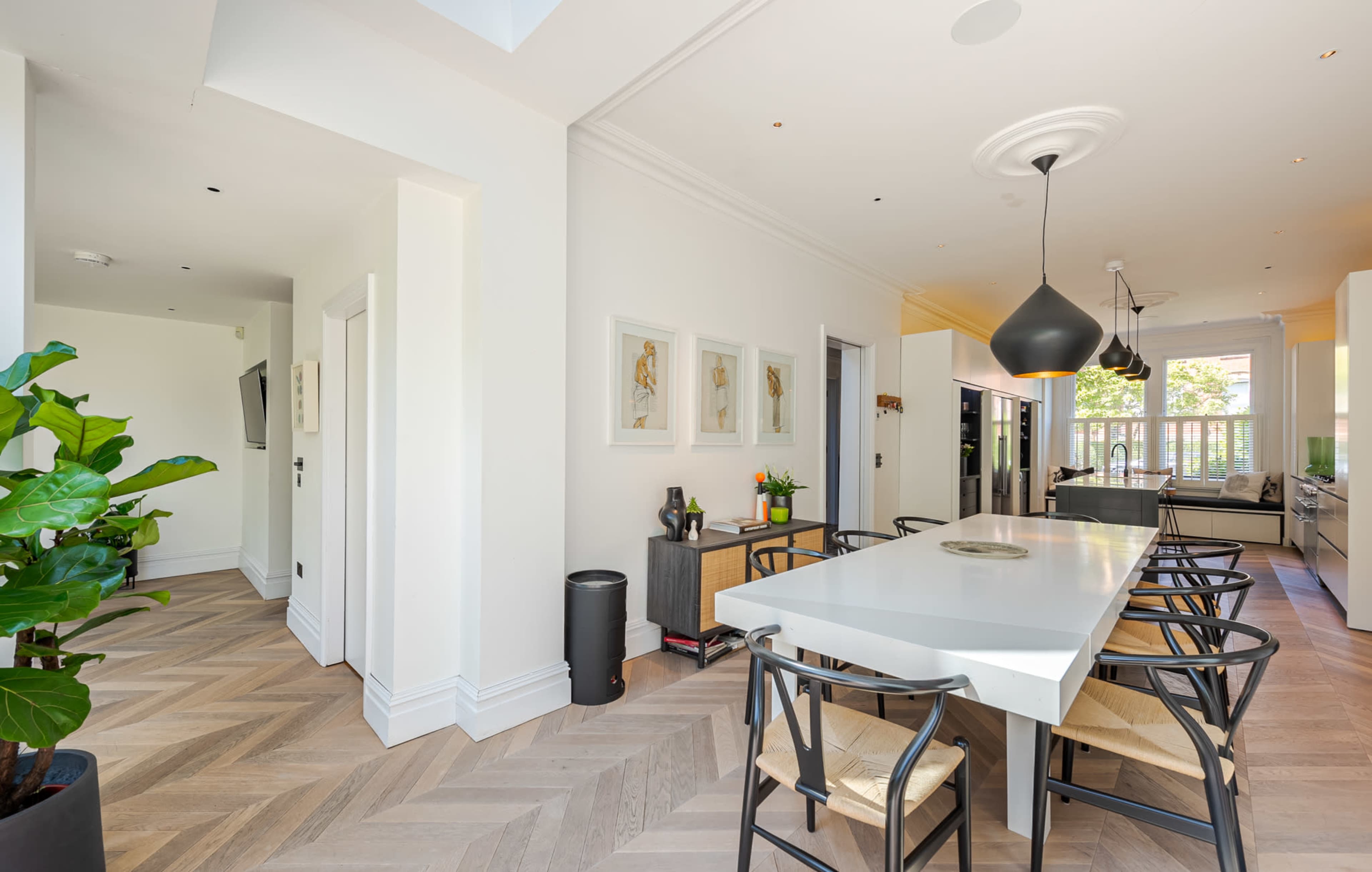 A modern dining area features a long table with black chairs, light hardwood flooring, and large windows allowing natural light to fill the space.
