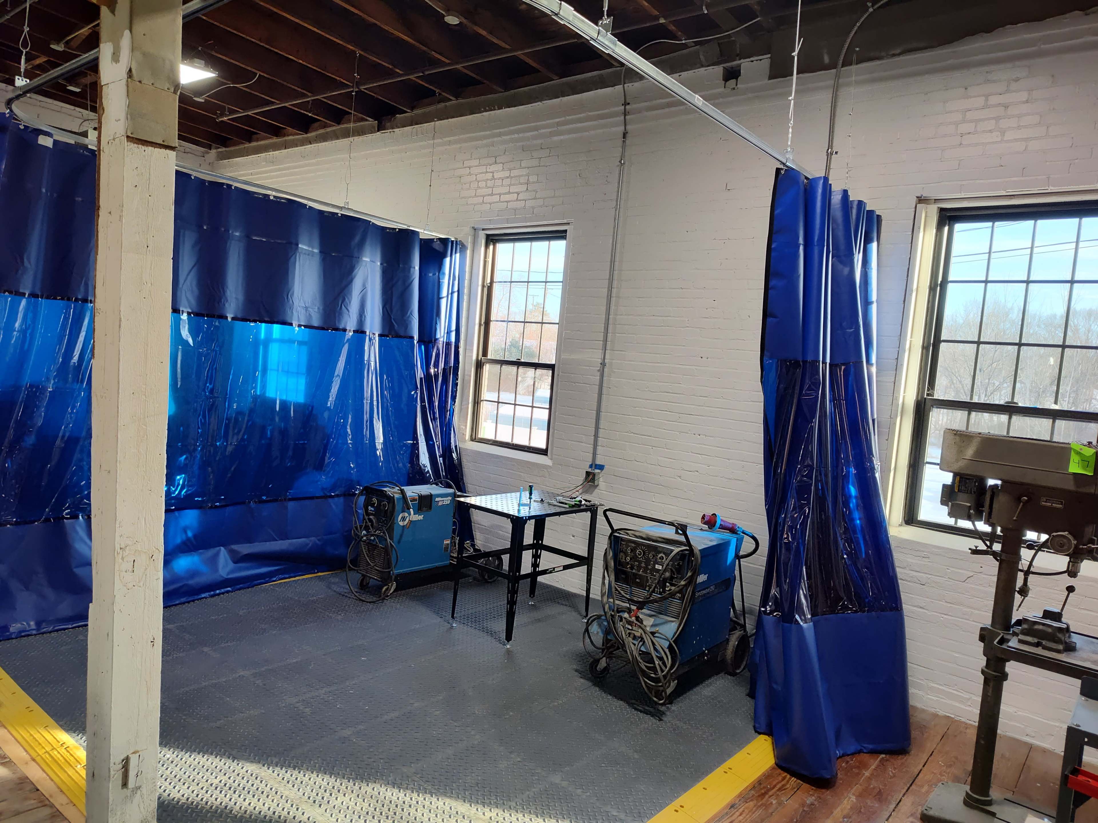 The image shows a work area enclosed with blue curtain panels, containing two welding machines and a table, situated near windows that allow natural light to enter.