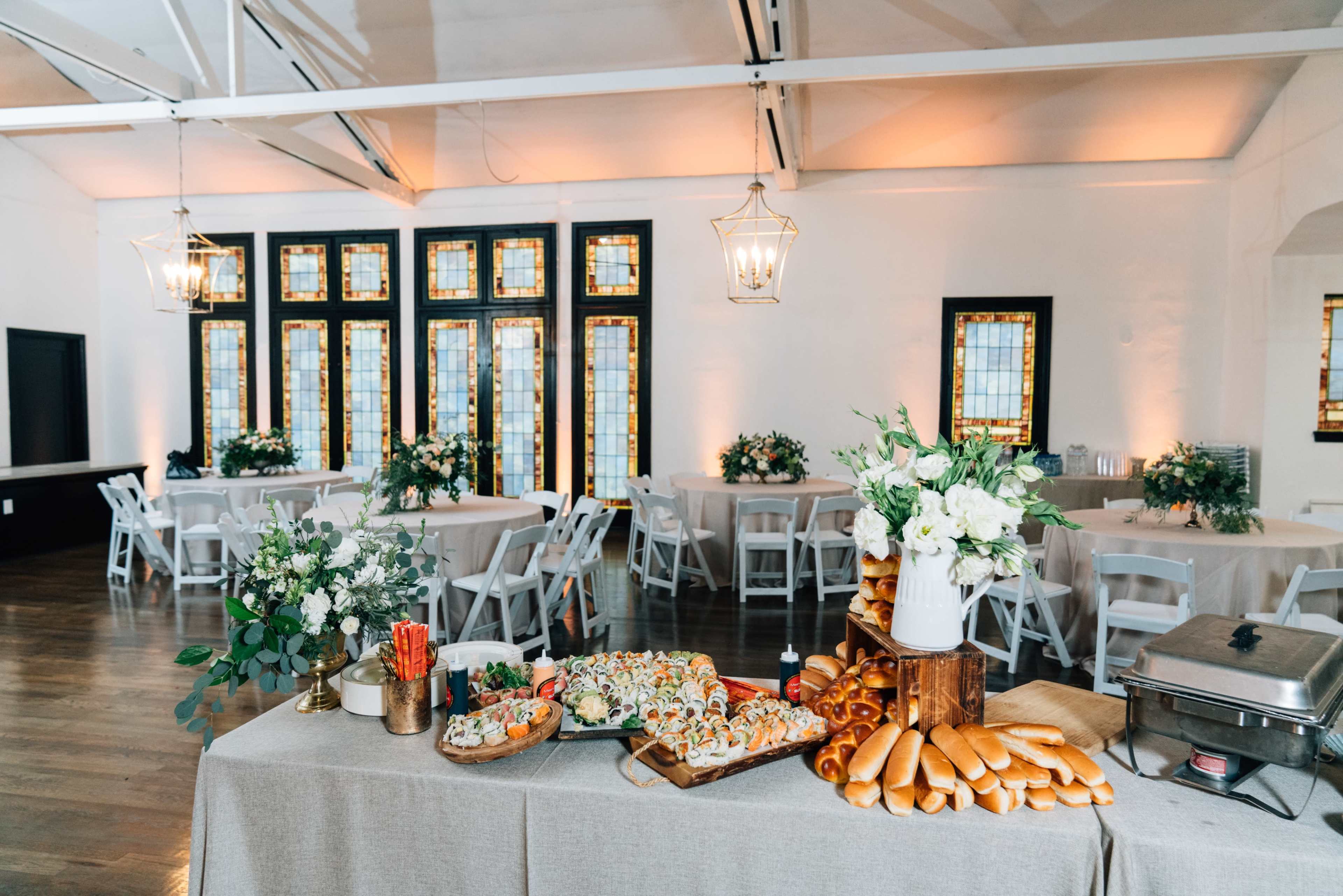 A beautifully arranged catering table displays a variety of food items, including sushi and bread, in a well-lit event space with round tables set for guests.