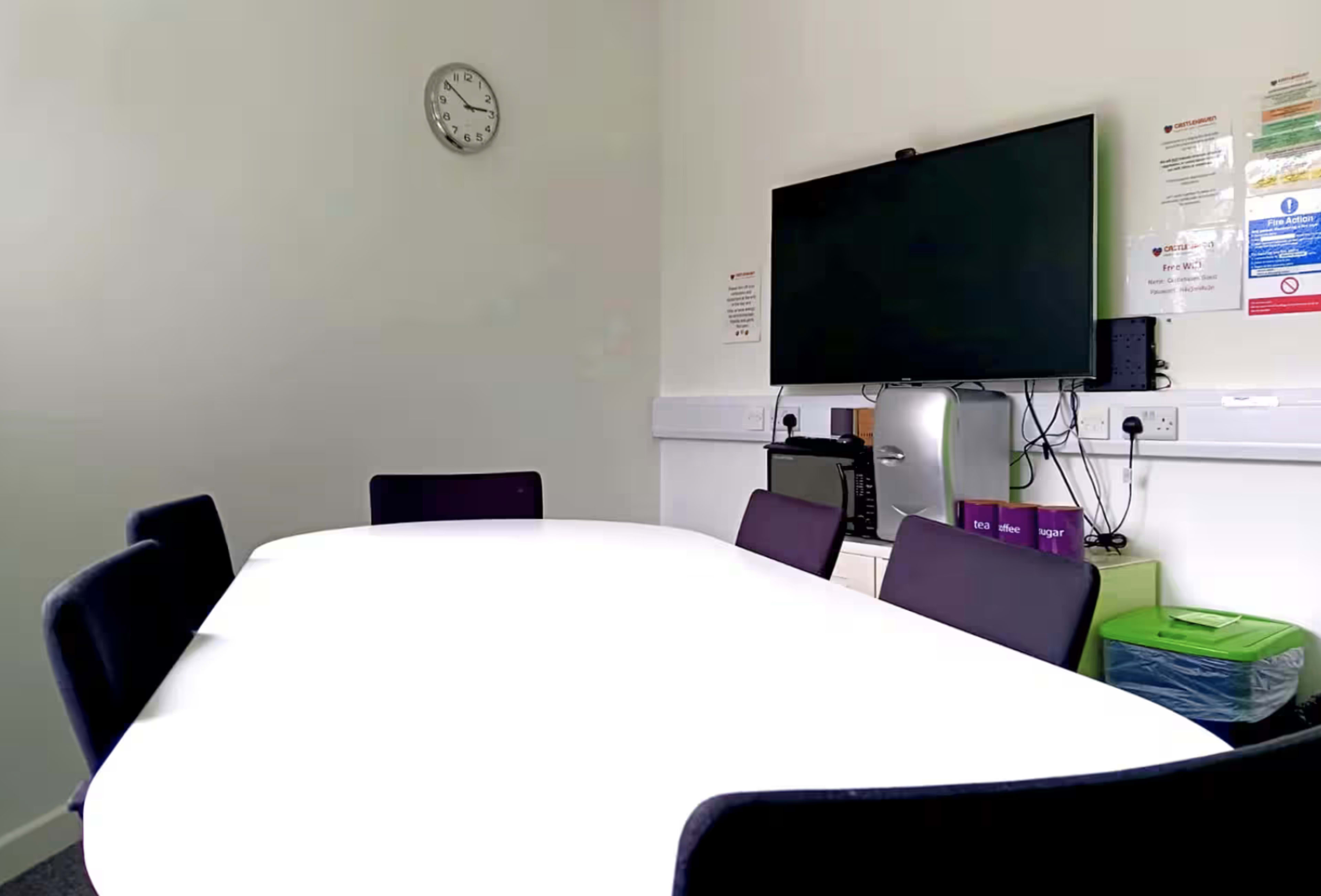 A conference room features a large white table, several purple chairs, a clock on the wall, and a television mounted on the opposite wall.