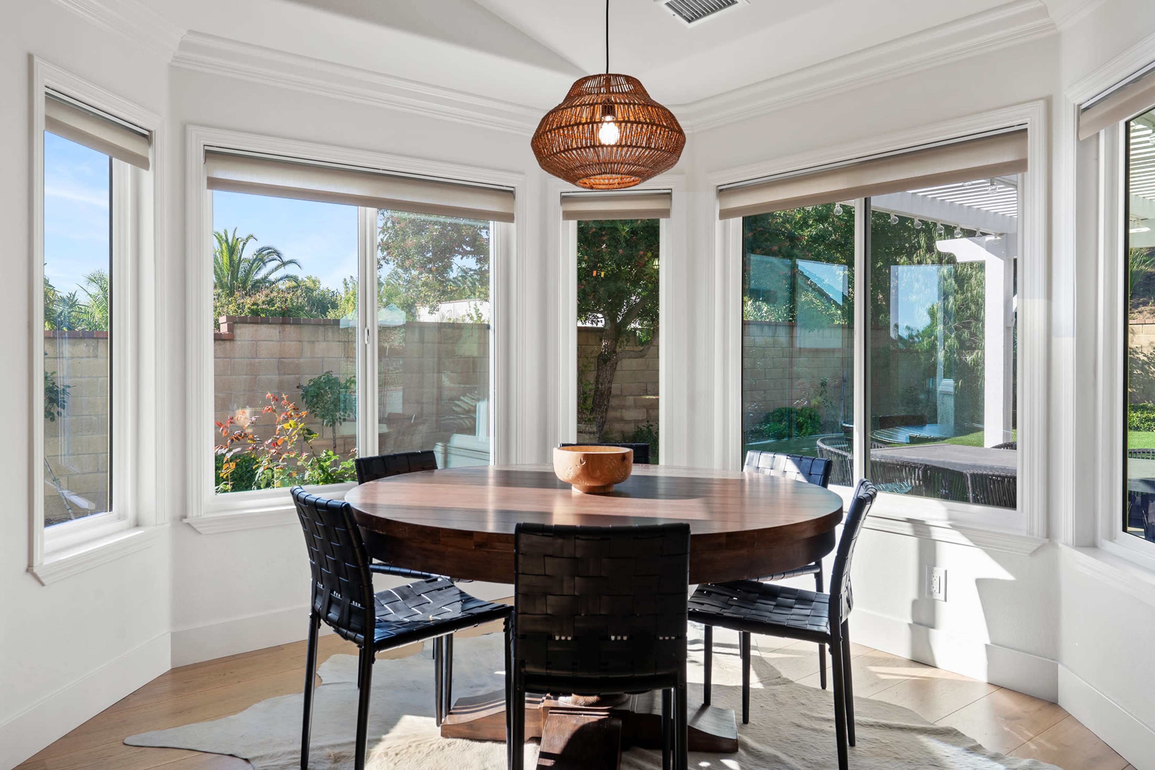 A round wooden table with black chairs is centered in a bright dining area surrounded by large windows showcasing a garden view.