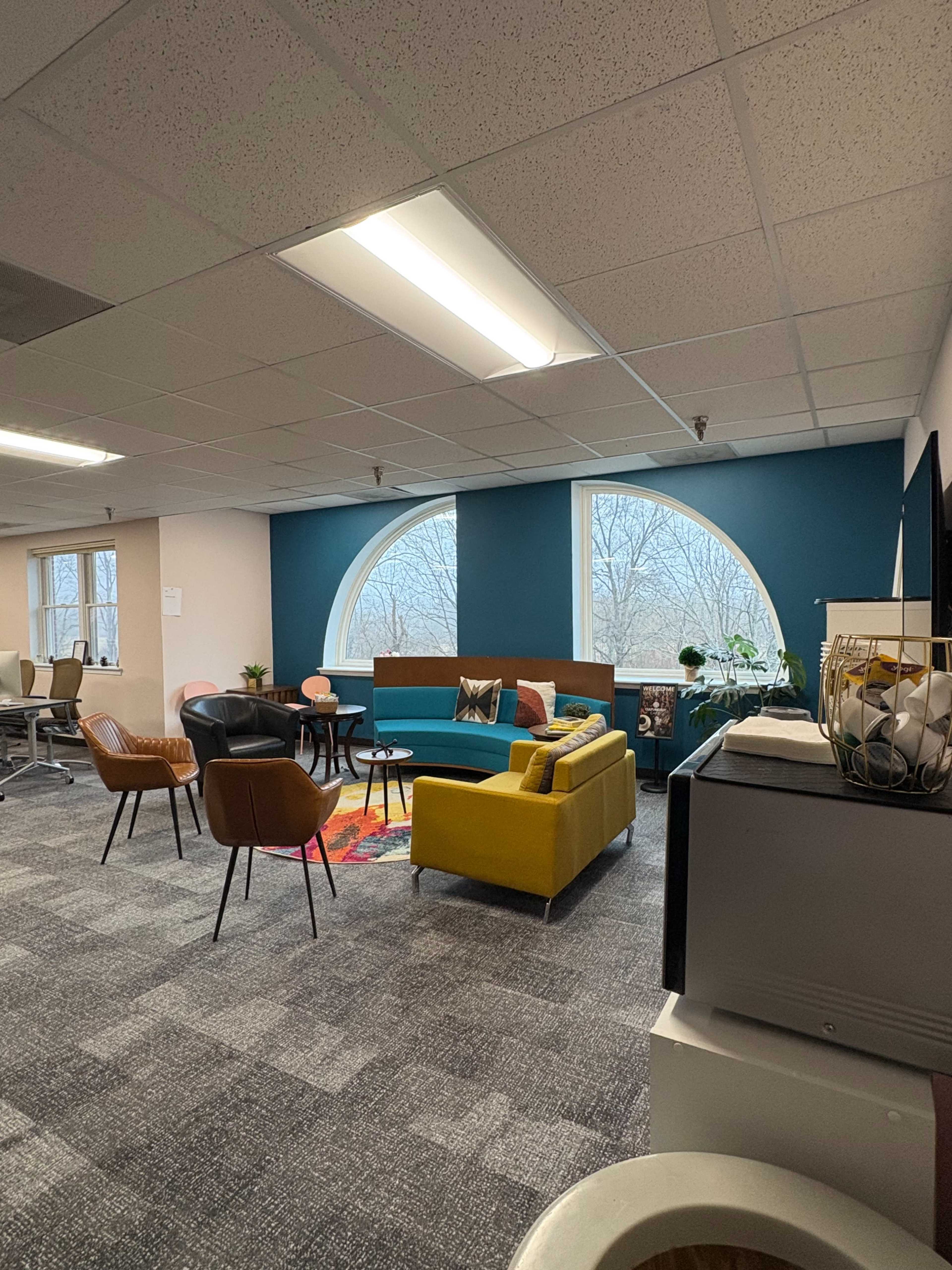 The image shows a cozy office lounge area with a large circular window, colorful seating, and plants.