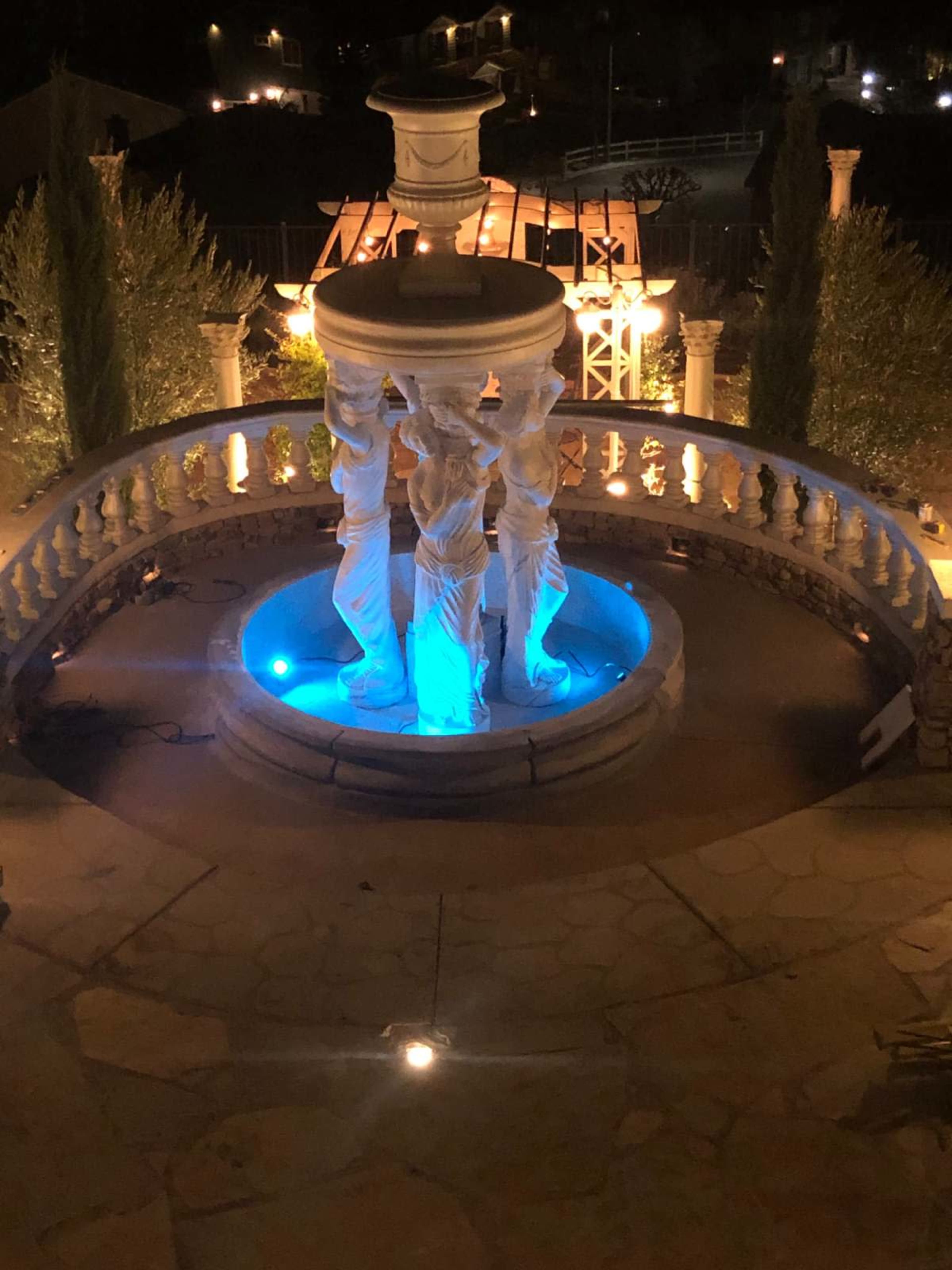 The image shows a marble fountain with four statues of women holding a basin, illuminated by blue lights, situated in a landscaped outdoor area at night.