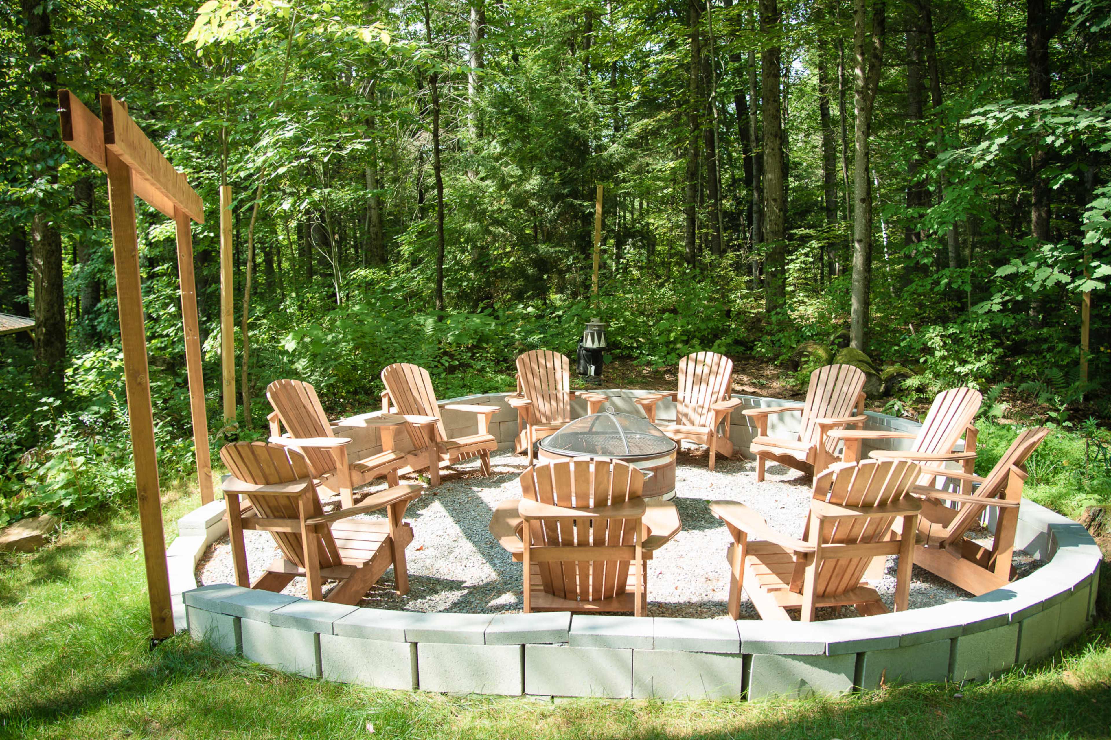 A circular patio area with eight wooden Adirondack chairs surrounds a stone fire pit, nestled in a wooded landscape.