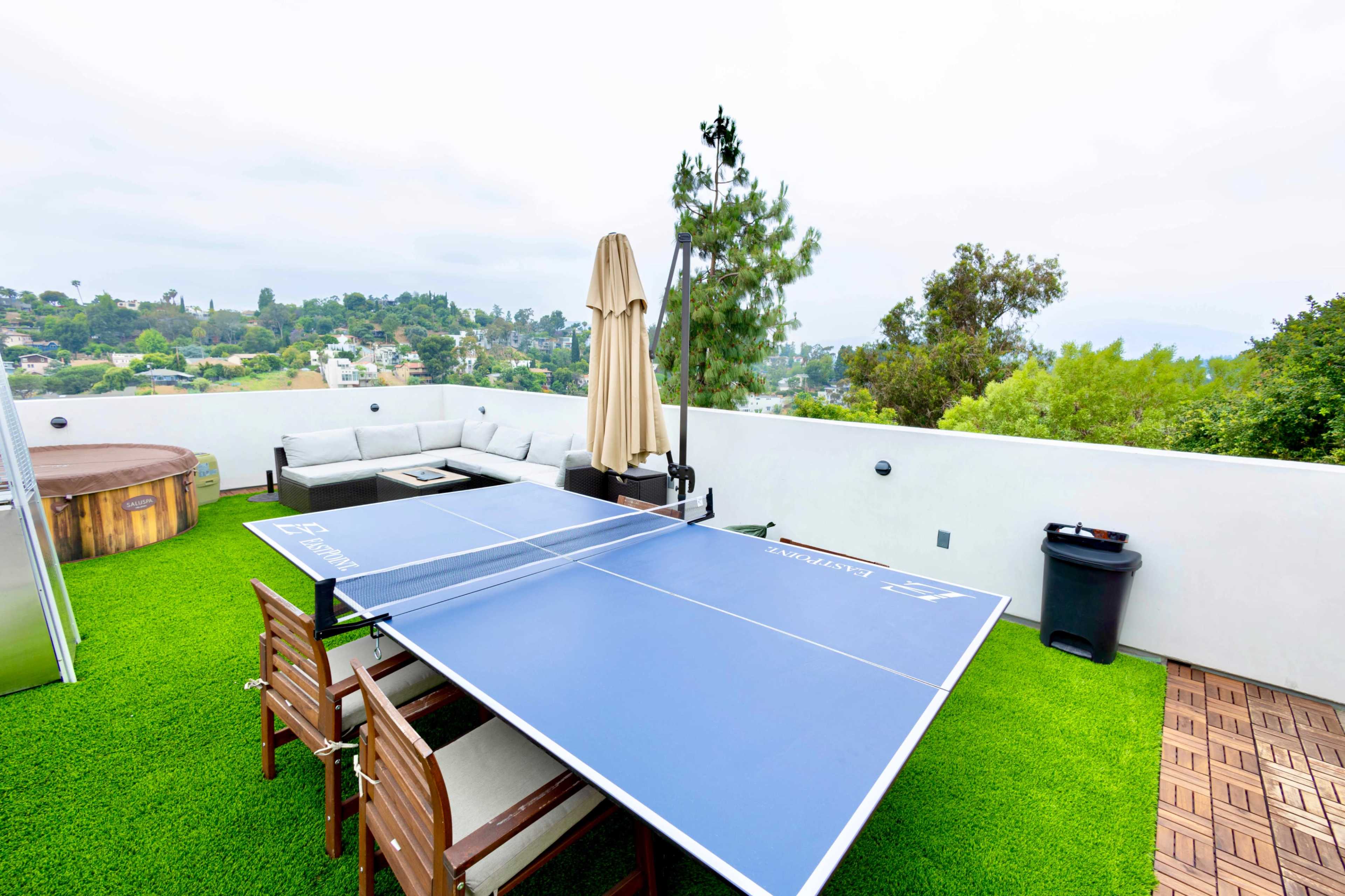 A rooftop space features a ping pong table, outdoor seating, a hot tub, and a patio umbrella surrounded by greenery.