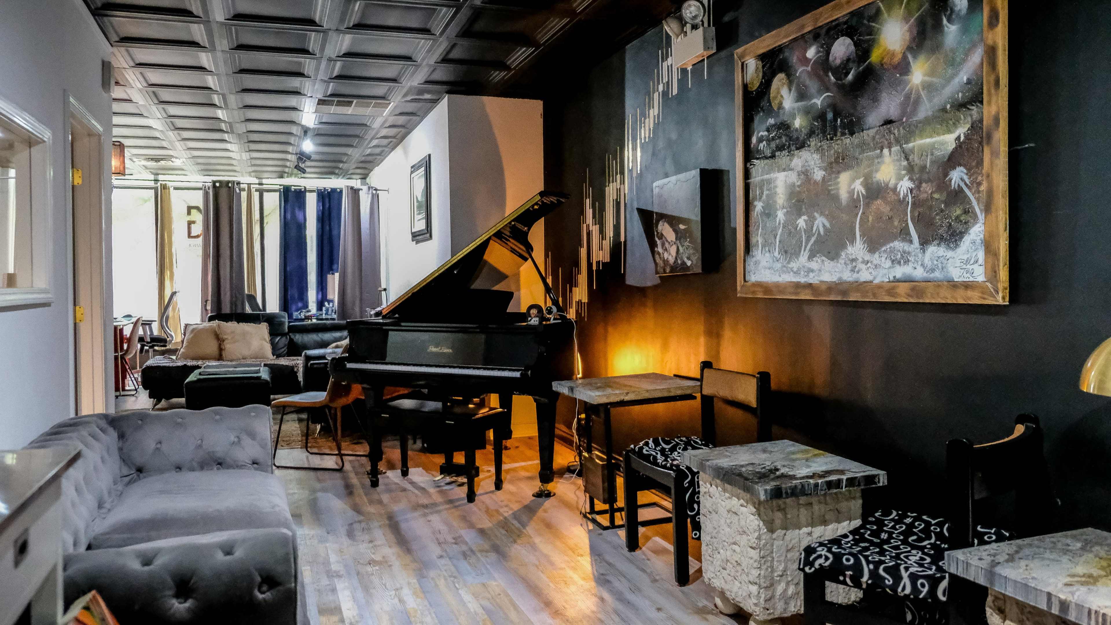 The image shows a stylish lounge area with a grand piano, various seating options, and artwork on the walls.
