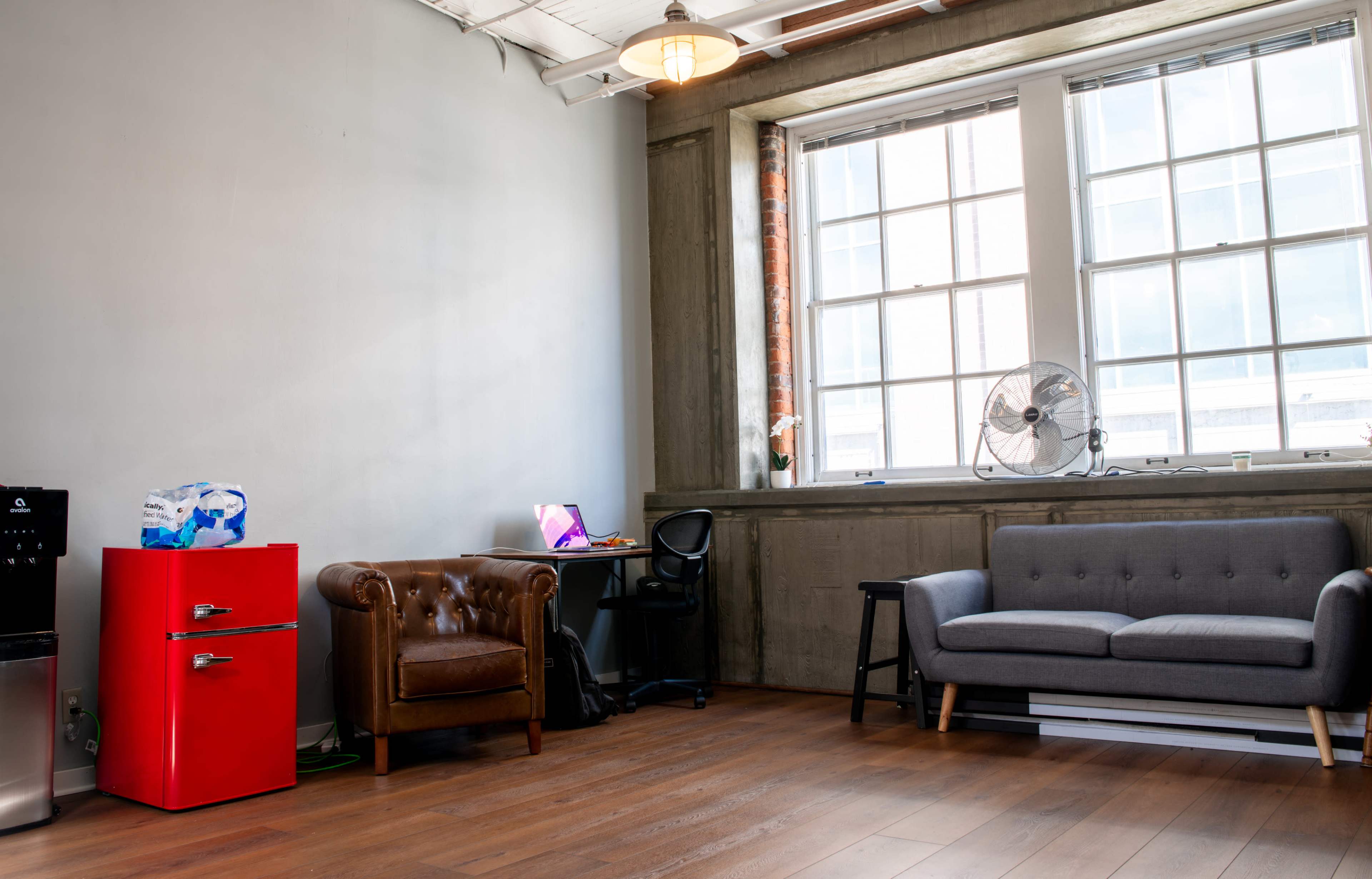 The image shows a modern office space with a red refrigerator, a brown leather couch, a desk with a computer, and large windows allowing natural light to enter.