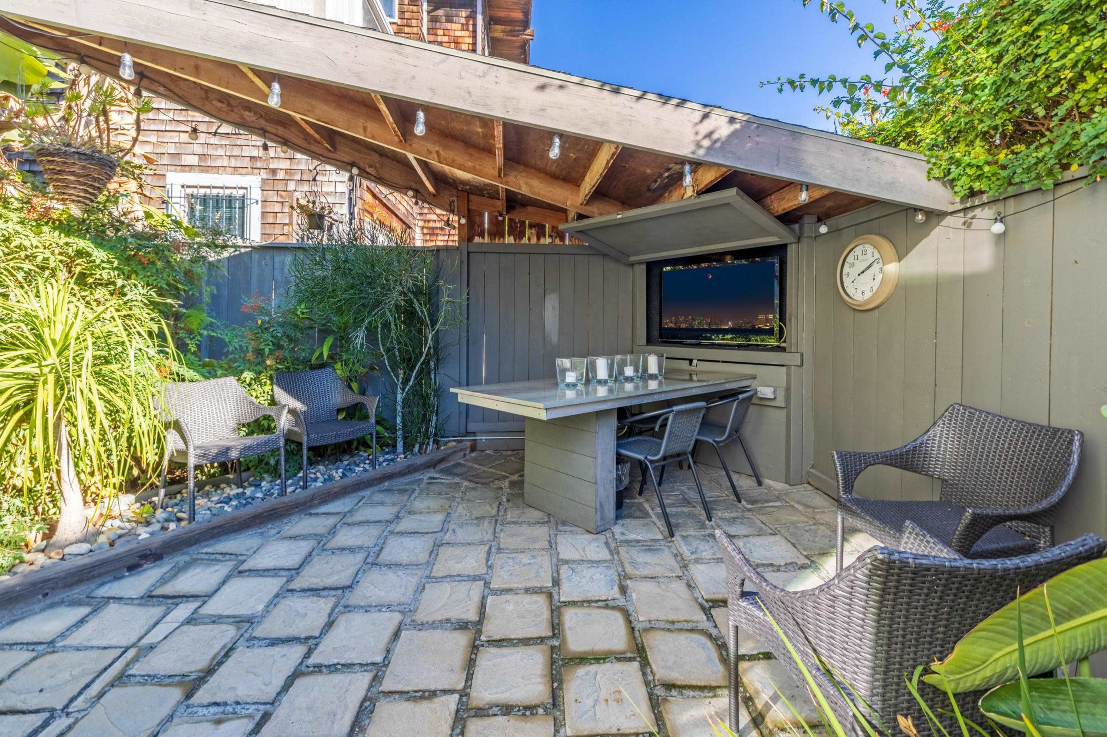 The image shows a covered outdoor seating area with a table and chairs surrounded by greenery and decorative stones.