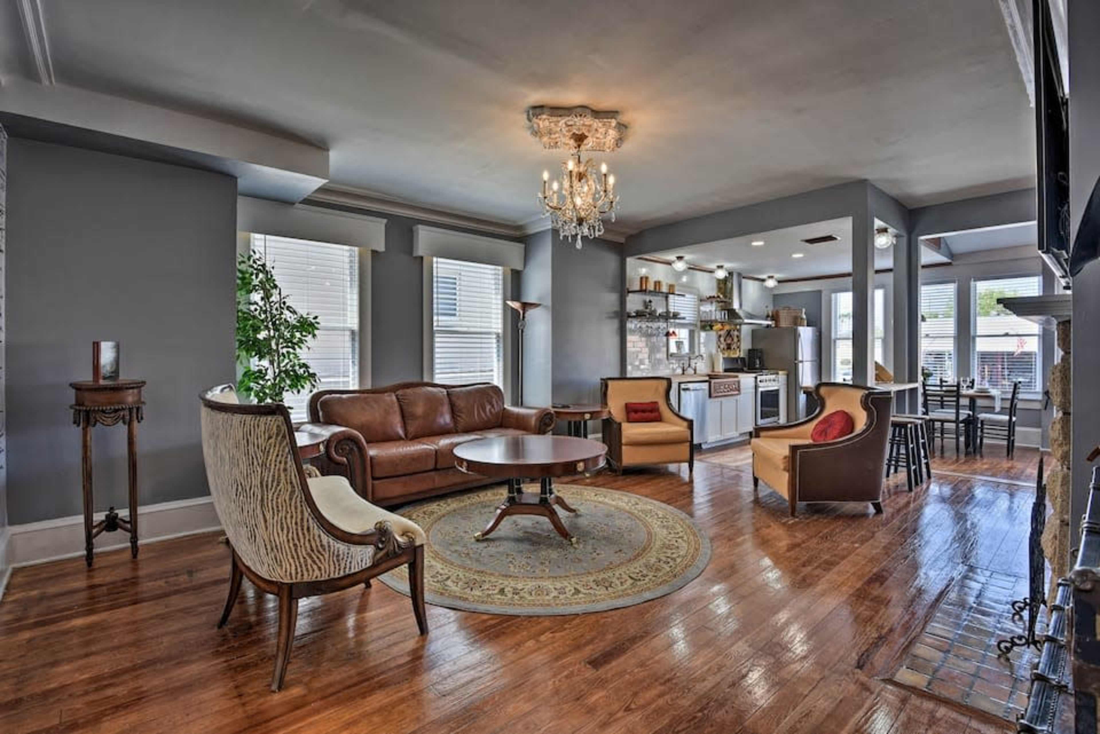 The image shows a spacious living room with hardwood floors, a brown leather sofa, and a round wooden coffee table, surrounded by windows and a kitchen area in the background.
