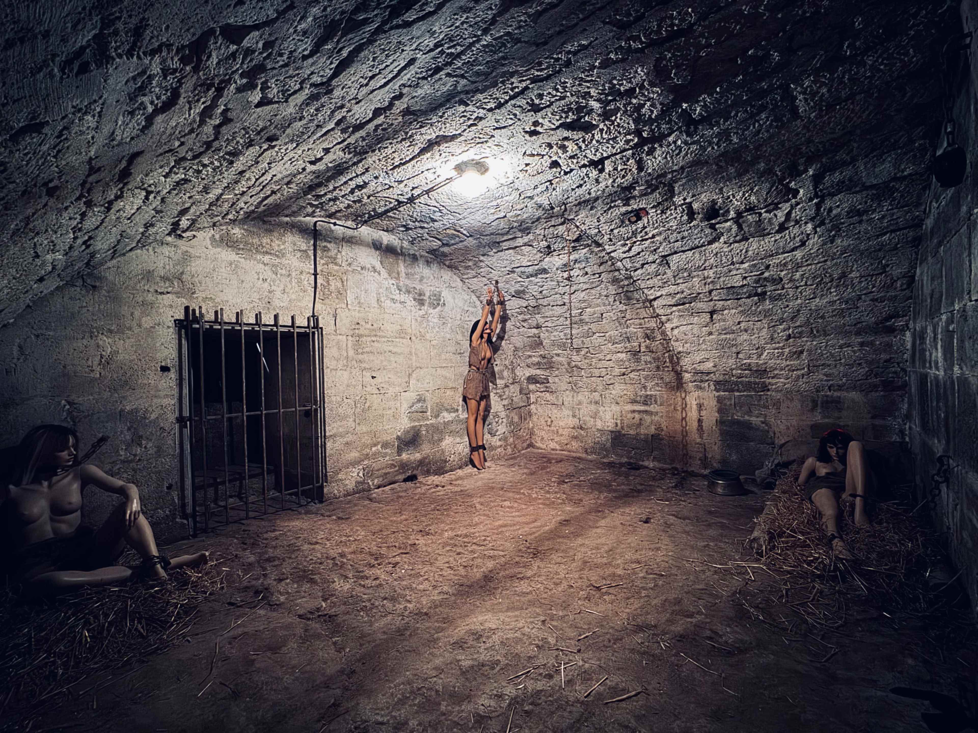 The image depicts a dimly lit, stone-walled dungeon with a person suspended from the ceiling and two others seated on the ground nearby.