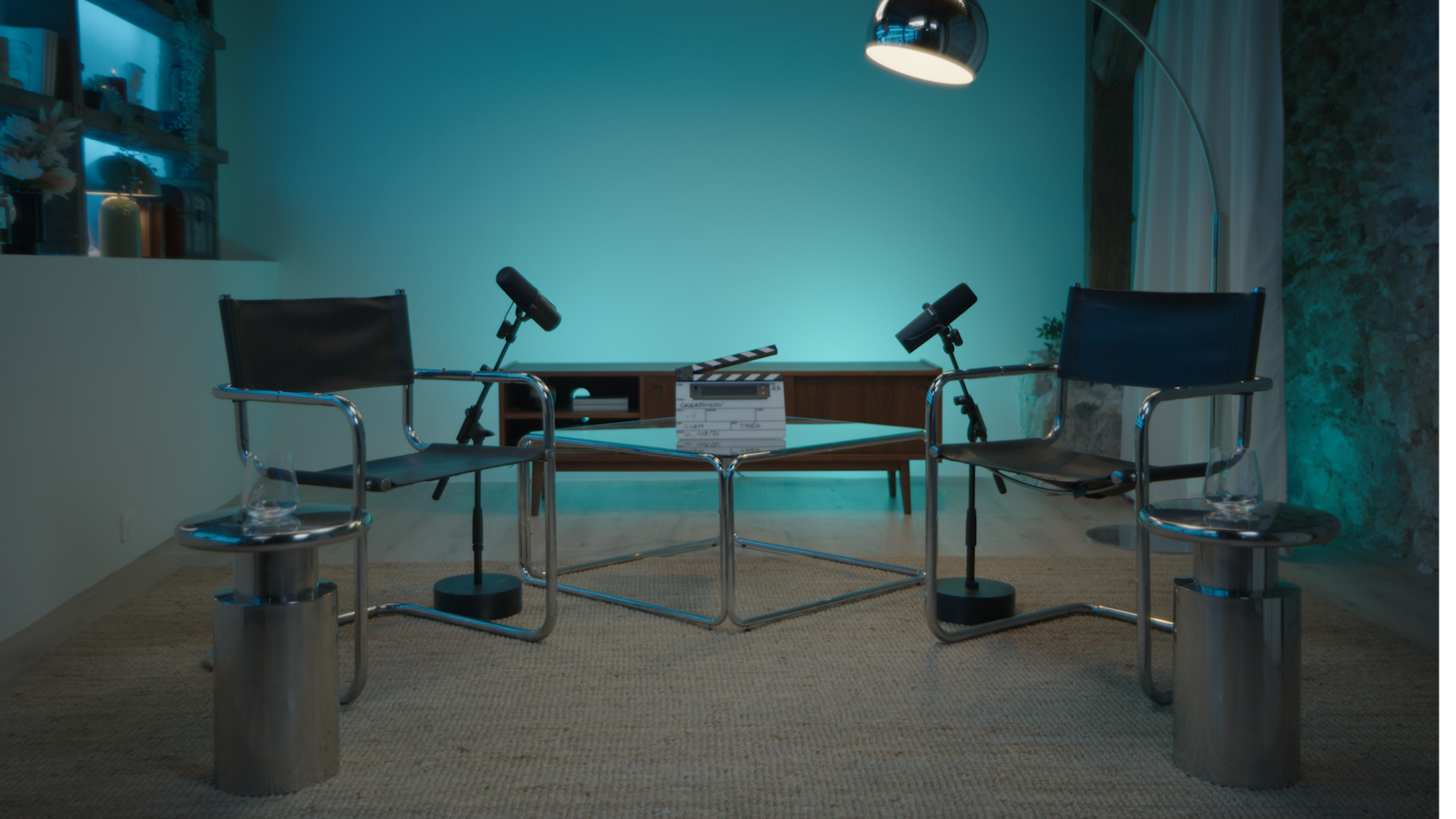 The image shows a minimalist interview setup with two empty chairs, microphones on stands, a coffee table with a clapperboard, and blue lighting in the background.