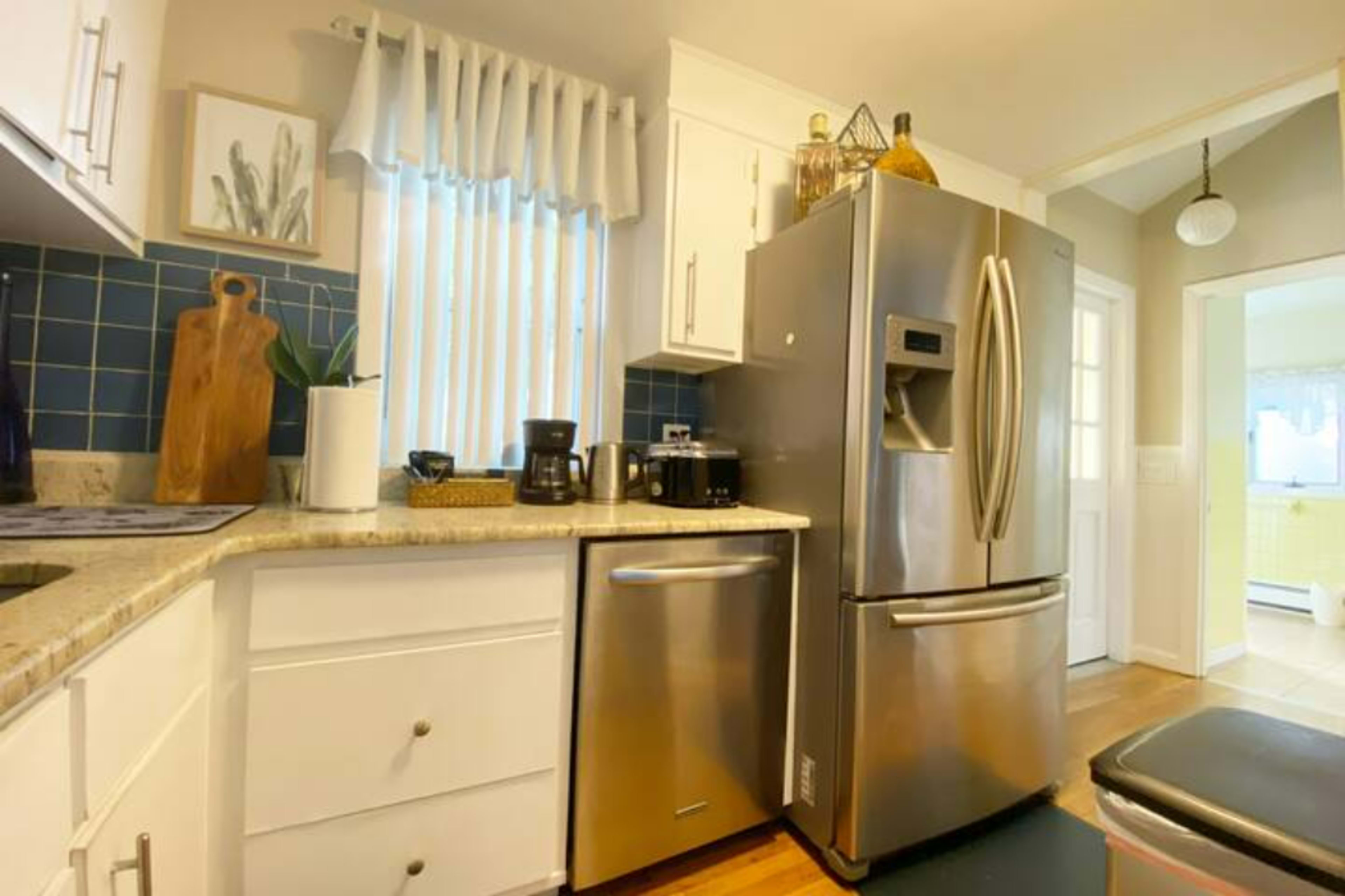 A kitchen with a stainless steel refrigerator, white cabinets, and a granite countertop, featuring a coffee maker and decorative items on the shelves.