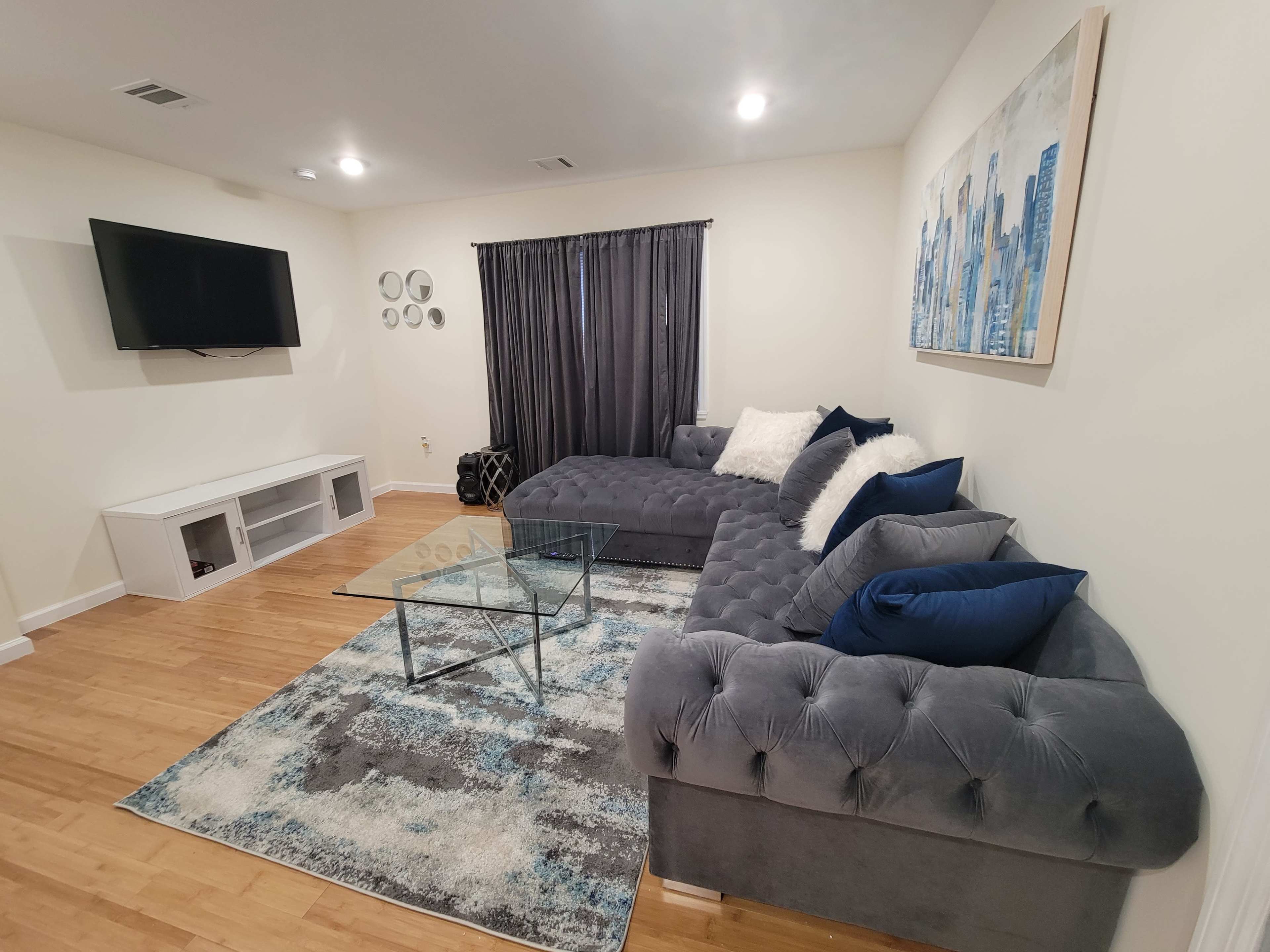 The living room features a gray sectional sofa, a glass coffee table, and a flat-screen TV mounted on the wall, with a large rug underfoot.