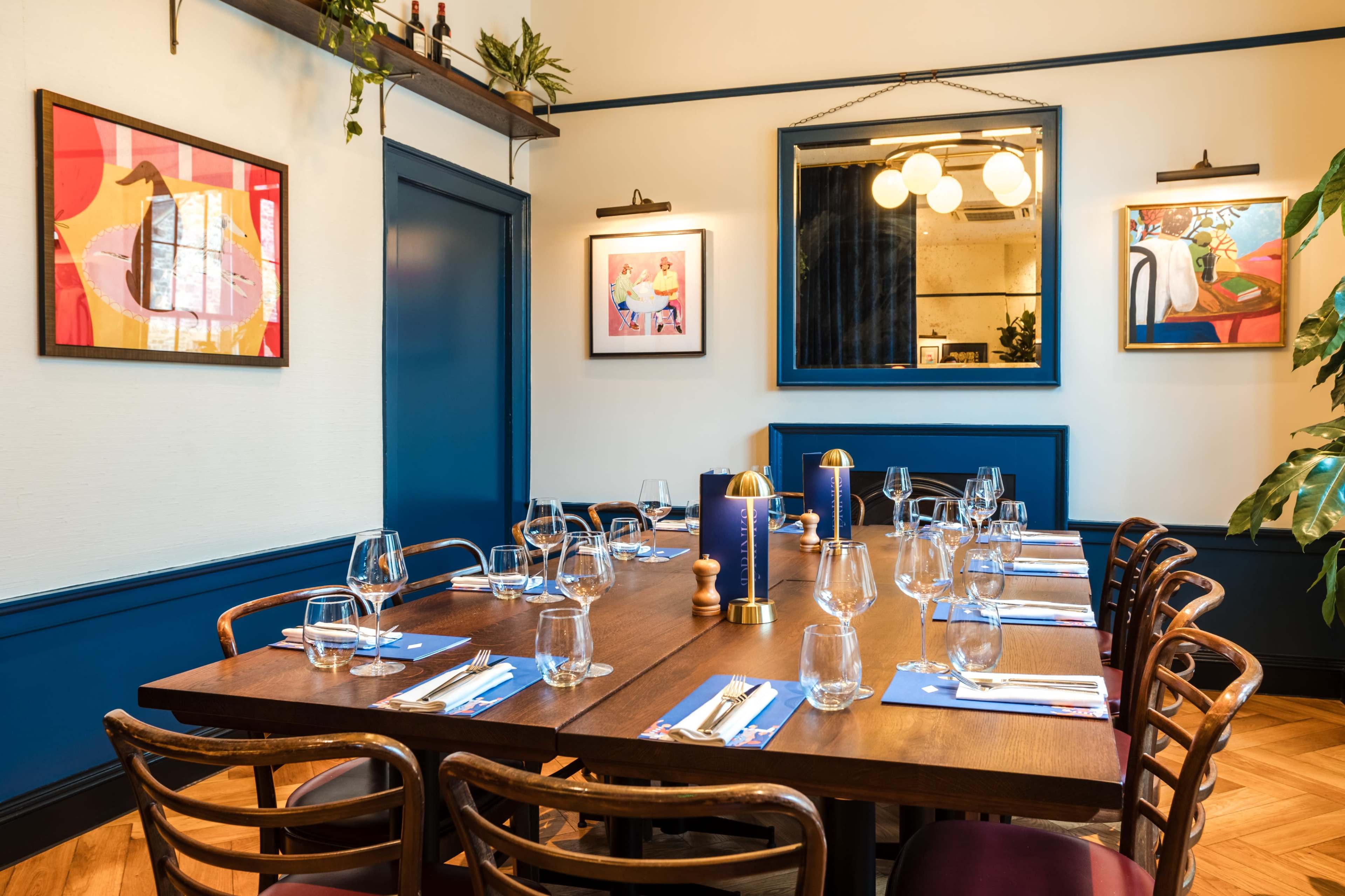 A dining room features a long wooden table set with glasses and silverware, surrounded by wooden chairs, with framed artworks on the walls and a large mirror reflecting the space.