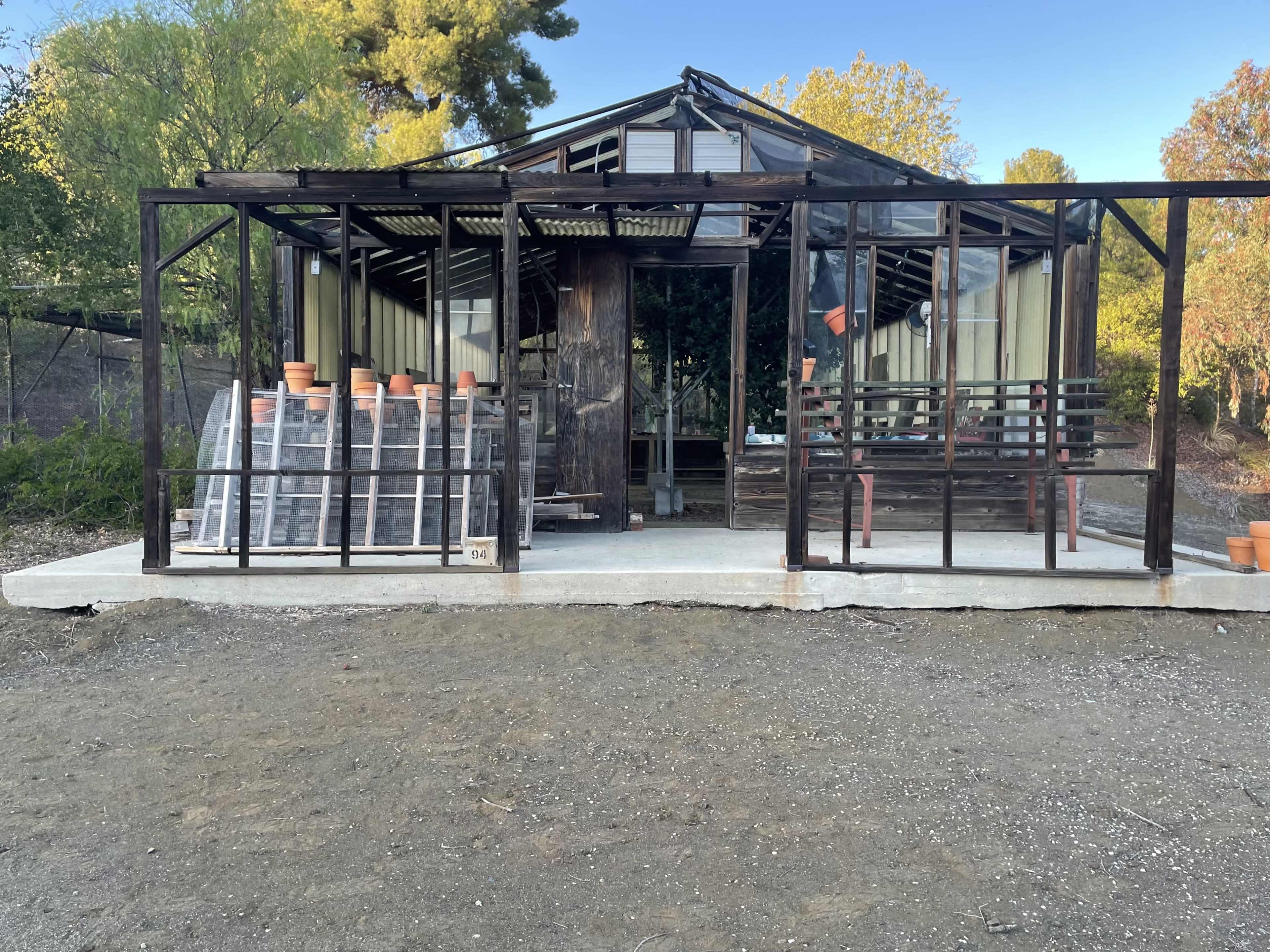 The image shows a greenhouse structure with glass panels, a wooden support post, and stacked pots outside on a gravel surface.